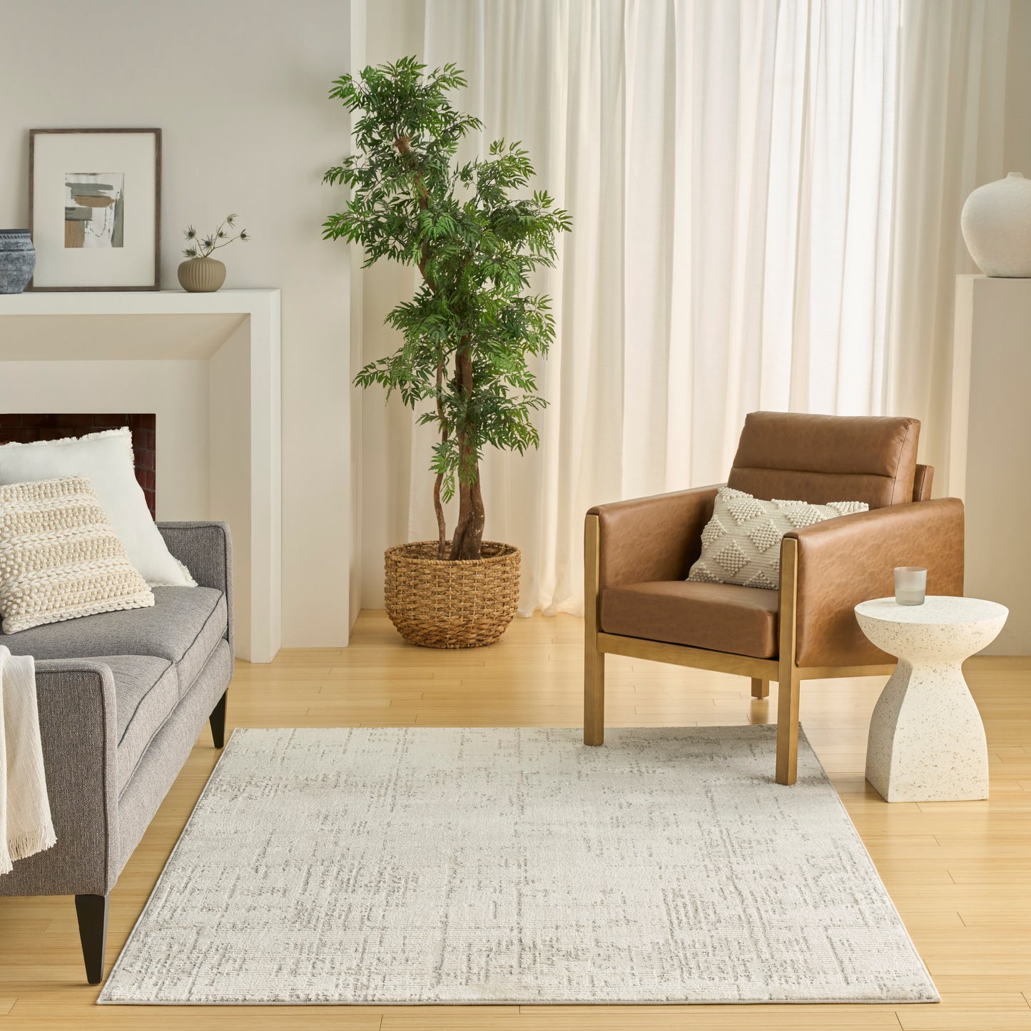 Neutral living room with gray fabric sofa, brown leather armchair, light rug, tall plant in woven basket, and white curtains.