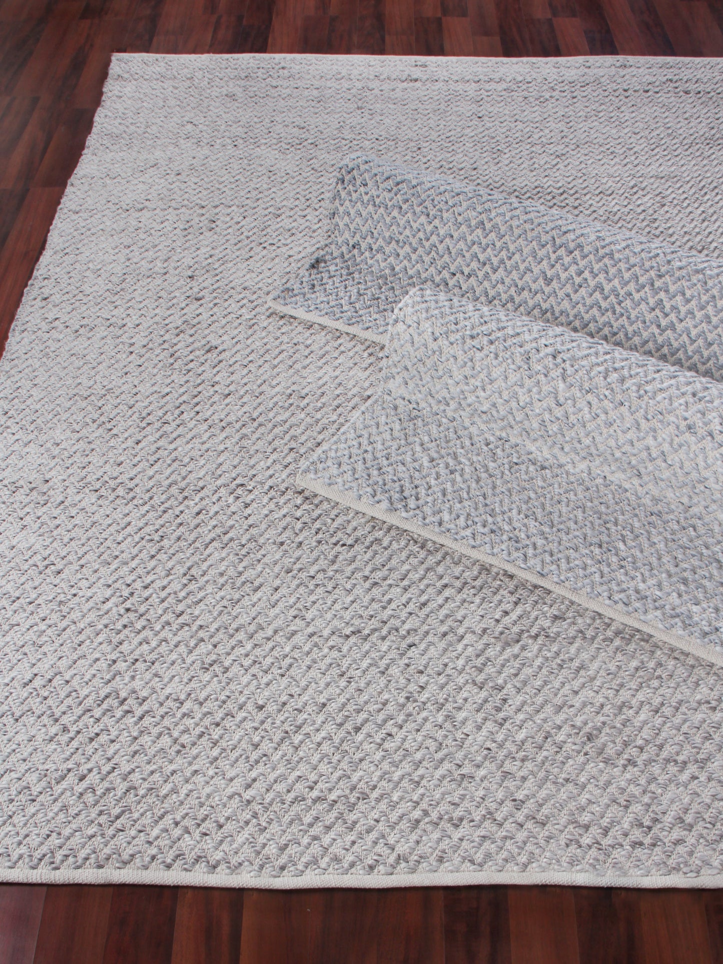 Light gray and white chevron woven rug on dark wood floor, textured loop weave with rolled corner.
