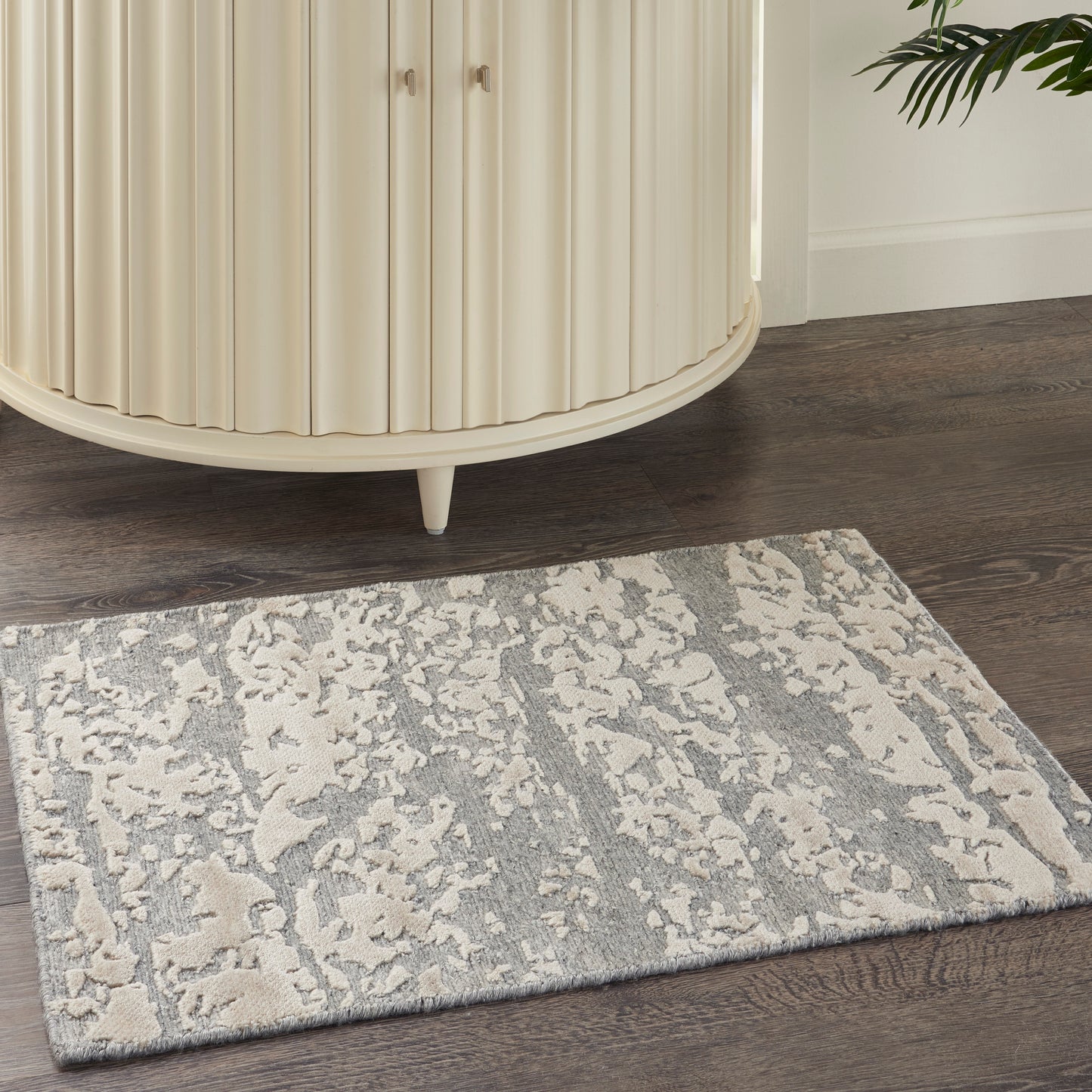 Beige curved ribbed cabinet on dark wood floor beside a gray patterned rug; plant peeks in.