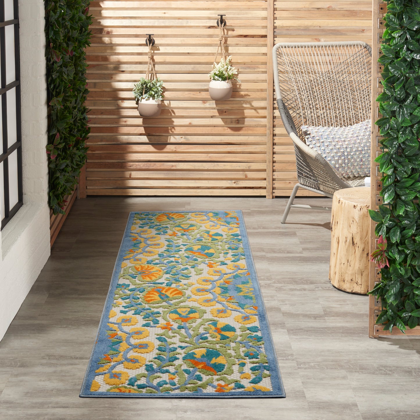 Outdoor runner rug with blue border and yellow-teal floral pattern on a wooden patio beside a wicker chair.
