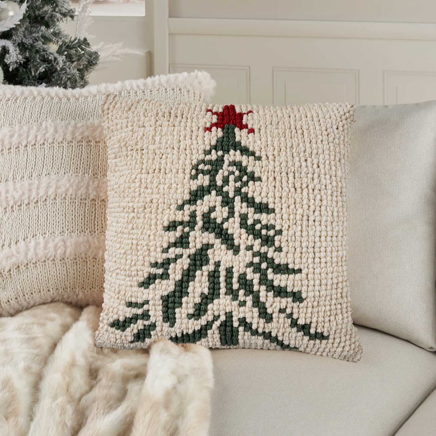 Cream chunky-knit decorative pillow featuring a green Christmas tree and red star, on a light sofa.