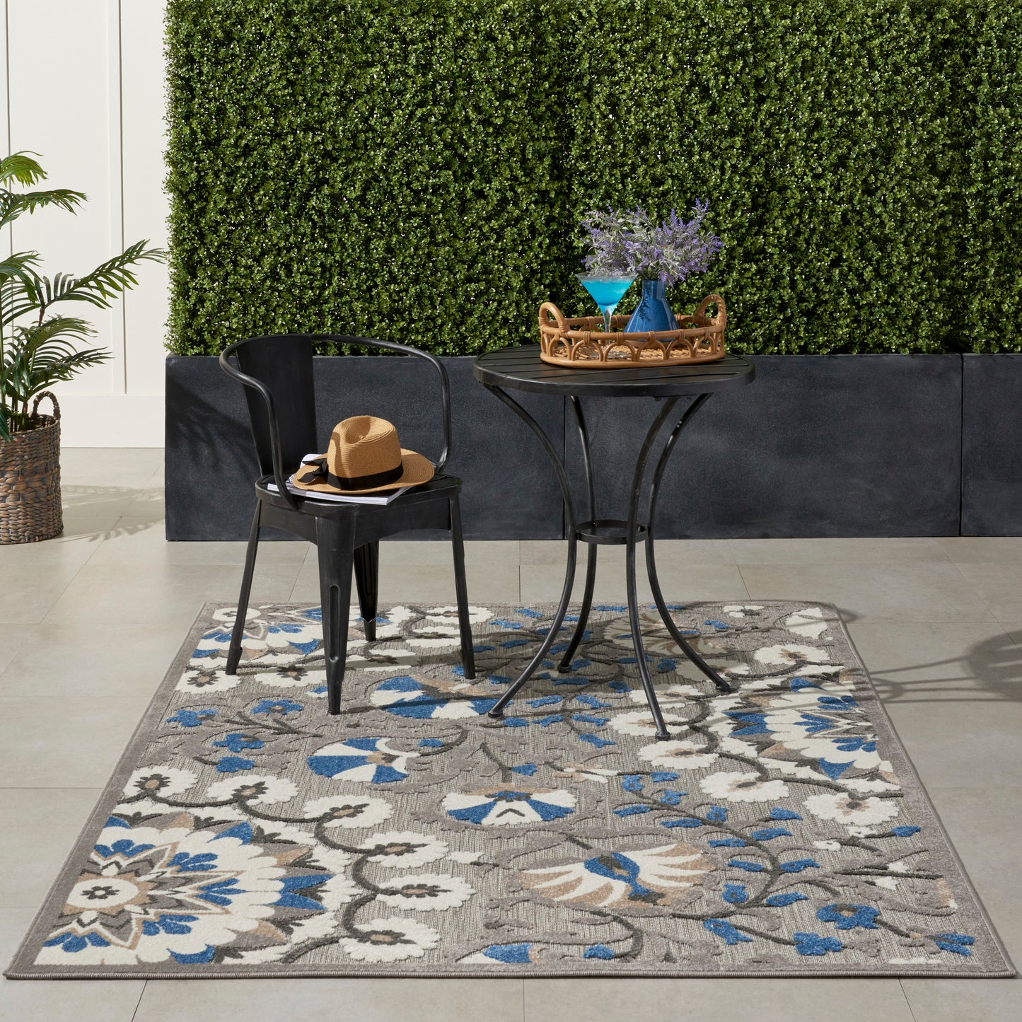 Outdoor black metal bistro set on blue-grey floral rug; round table with wicker tray and purple flowers in a blue vase.
