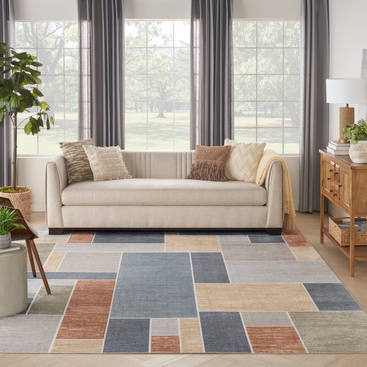 Colorful geometric patchwork rug on a living room floor, with a beige sofa and textured throw pillows, a potted plant, a wooden console, and dark drapes.