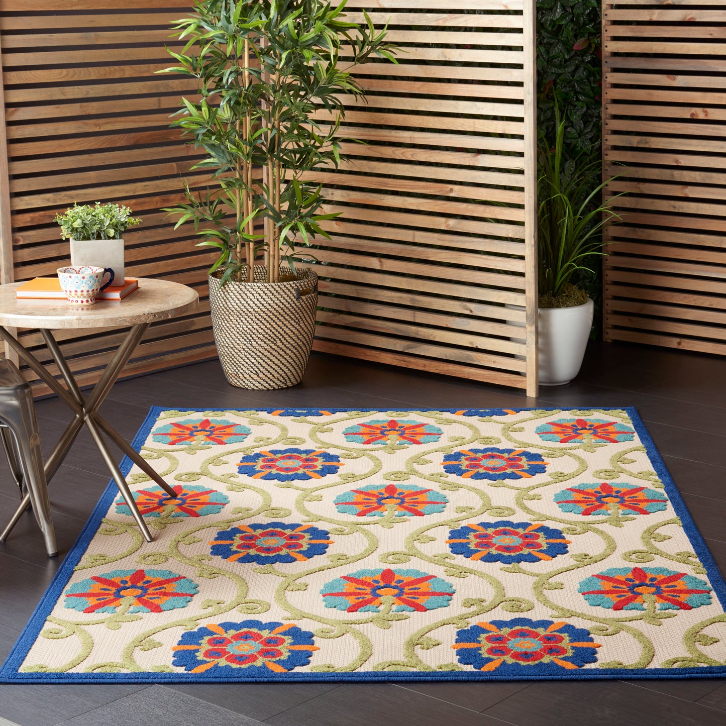 Beige outdoor area rug with blue border and repeating blue and red floral motifs, green vines, on a wooden deck.