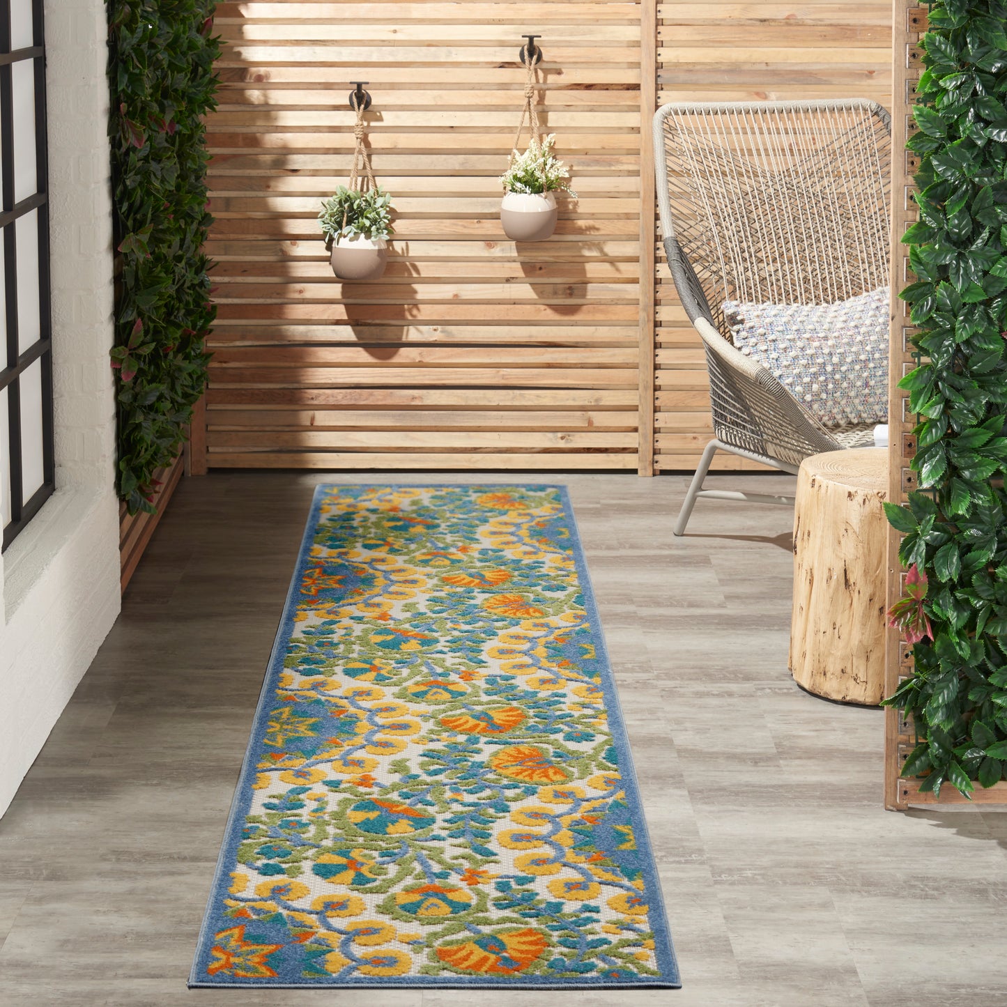 Outdoor runner rug with teal, yellow & orange floral print and blue border on a wooden deck beside a wicker chair.