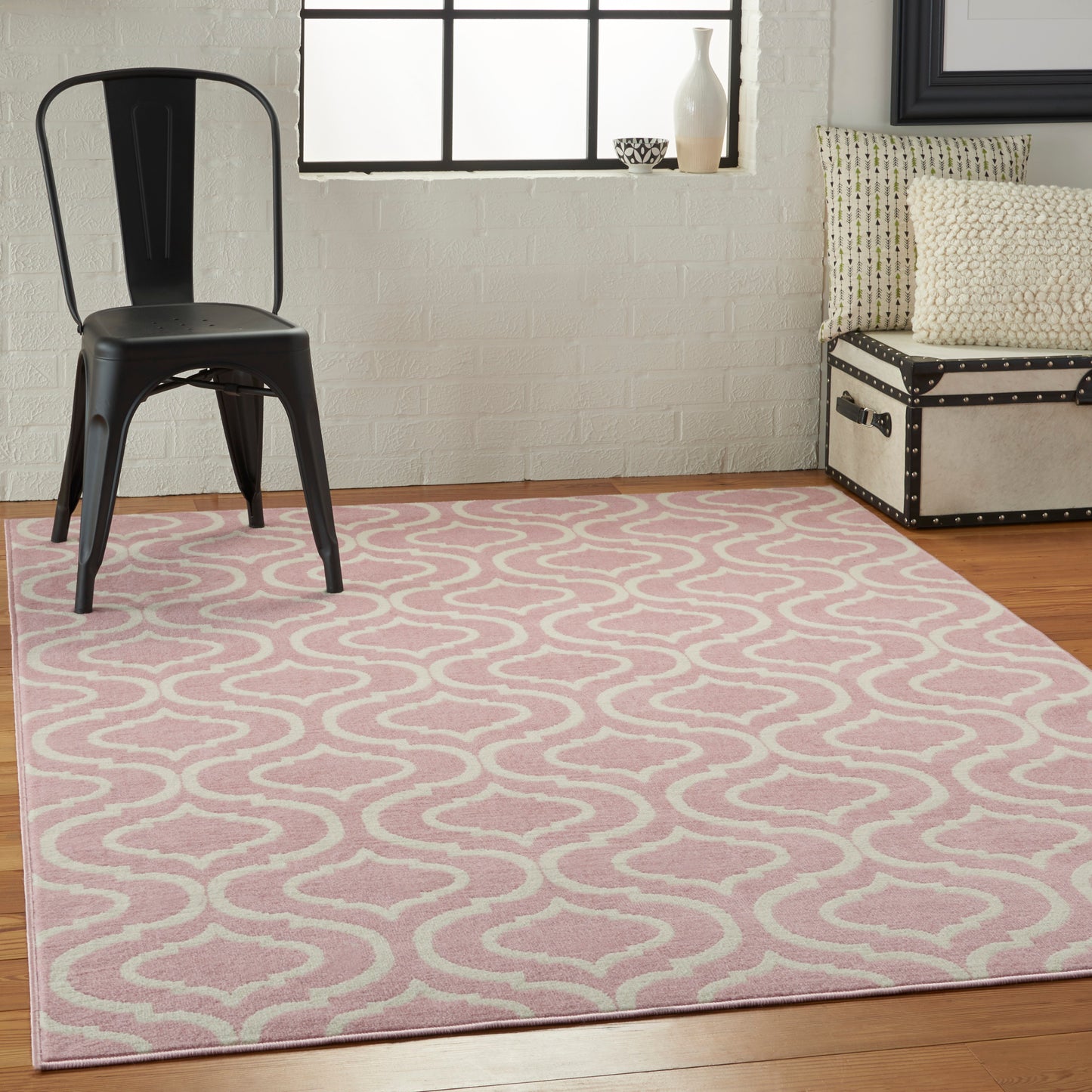 Pink wavy-pattern area rug on a wood floor with a black metal chair and a beige studded trunk with cushions.