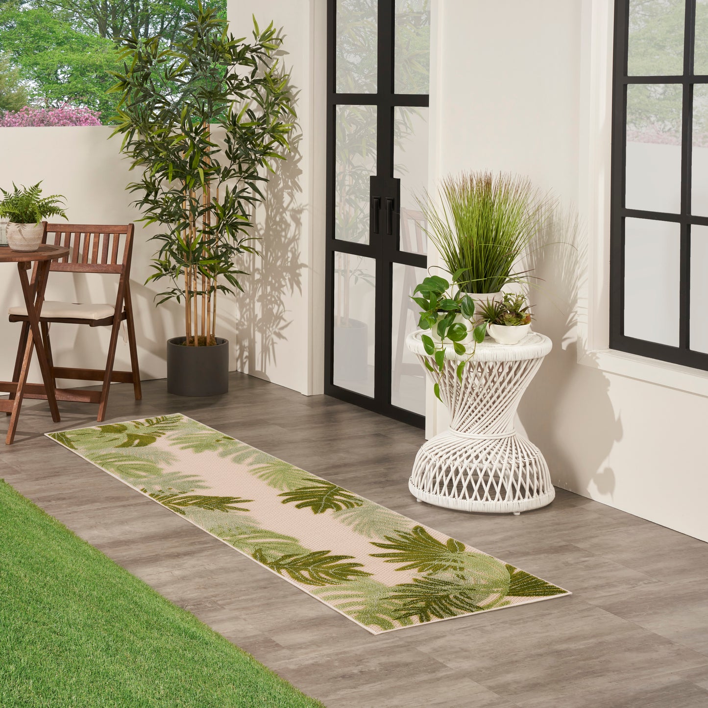 Long palm-leaf patterned beige-green runner rug on wood floor in a bright plant-filled room with black-framed doors.