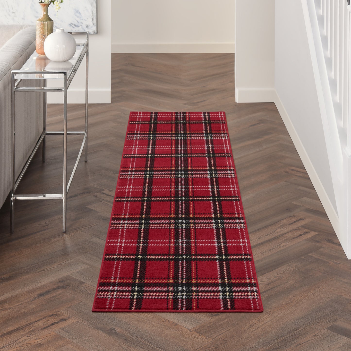 Red plaid tartan runner rug in a modern entry hall with dark wood herringbone floor and a metal-framed glass console table.