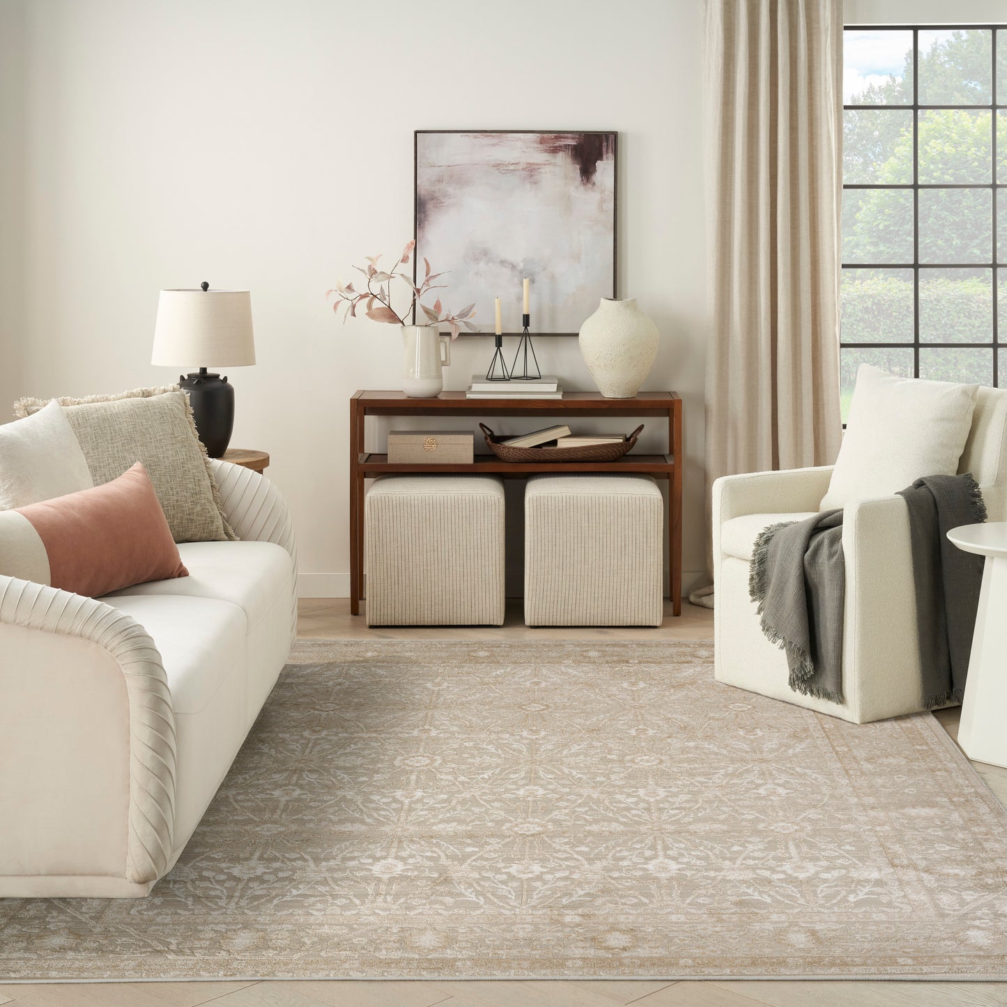 Neutral living room with cream sofa, blush pillow, wooden console, two beige ottomans, armchair, and light patterned rug.