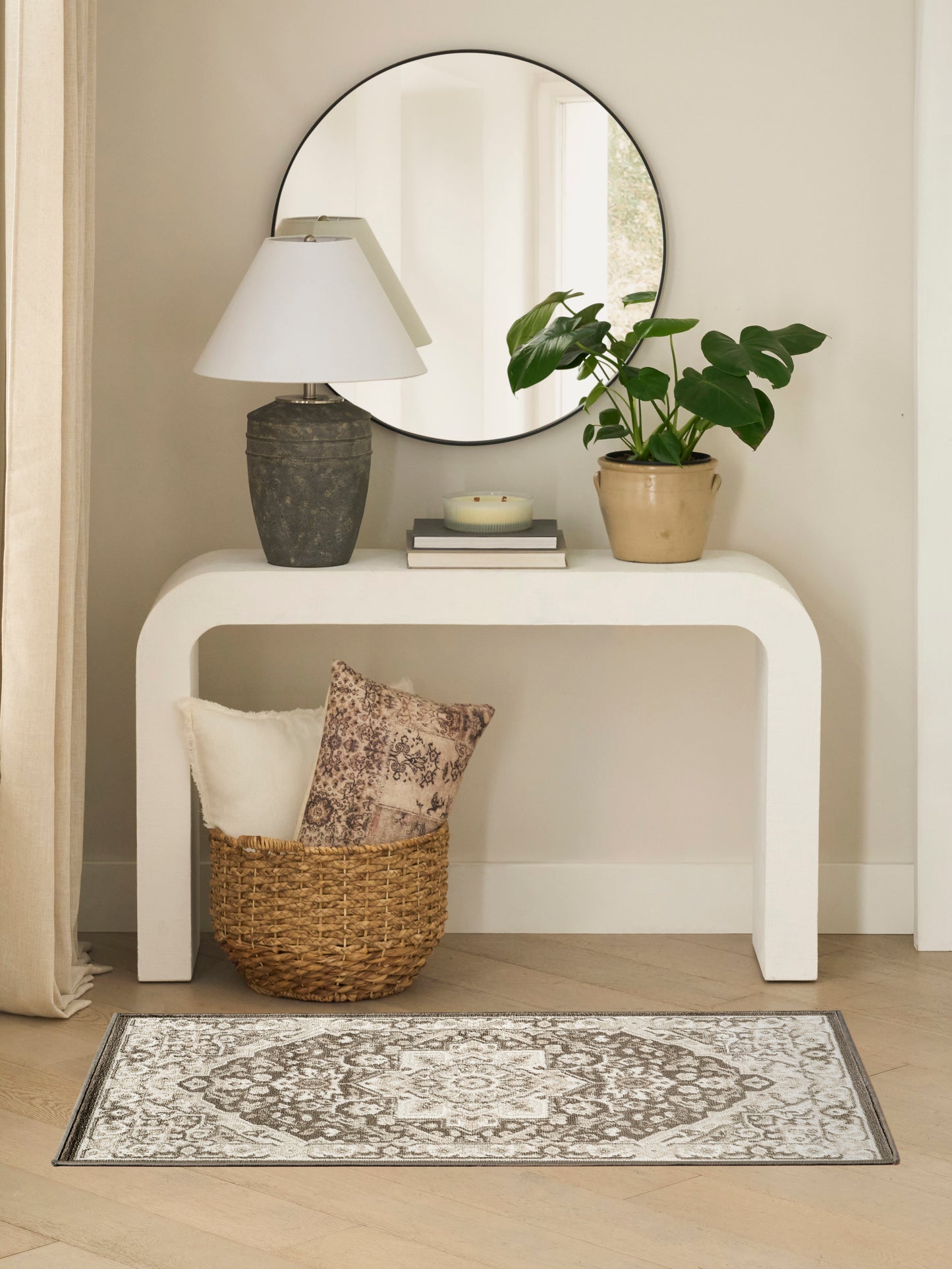 Round mirror above a white curved console with a stone lamp, potted plant, books and a woven basket on a gray patterned rug.