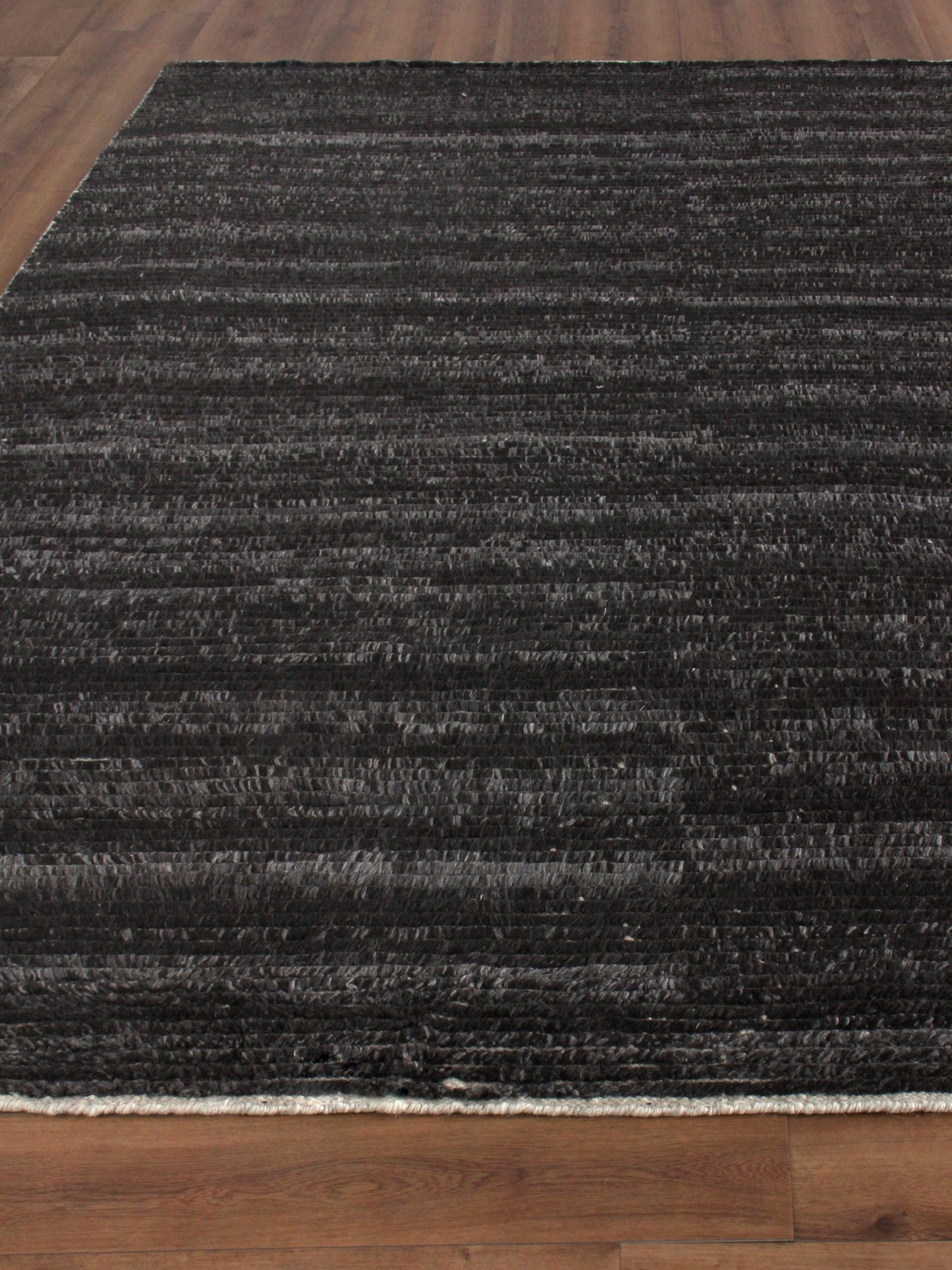 Dark charcoal rug with subtle variegated gray stripes, plush flat-weave, edge fringe on bottom, placed on wood floor.
