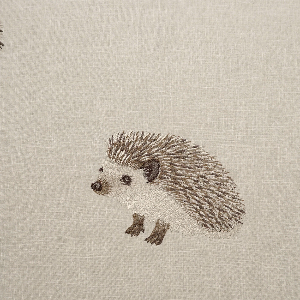 Embroidered hedgehog on beige linen fabric; cream body with brown quills and tiny feet.