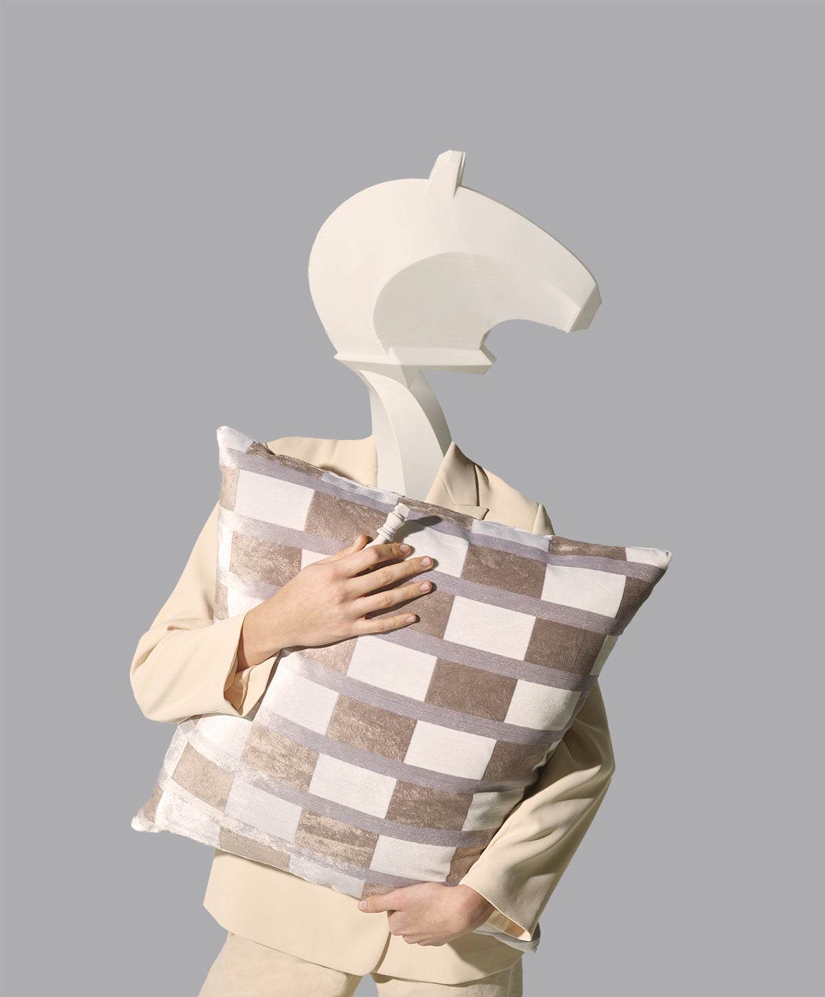 Beige-suited figure wearing a white abstract horse-head mask, holding a large beige-and-white checkered pillow.