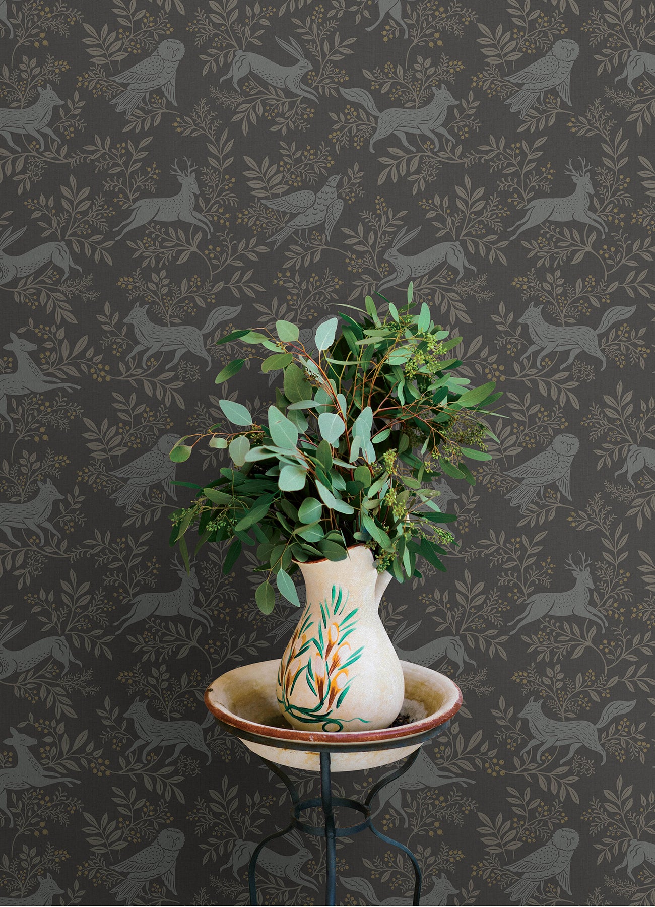 Hand-painted ceramic vase with green foliage bouquet on a metal stand against dark botanical wallpaper.