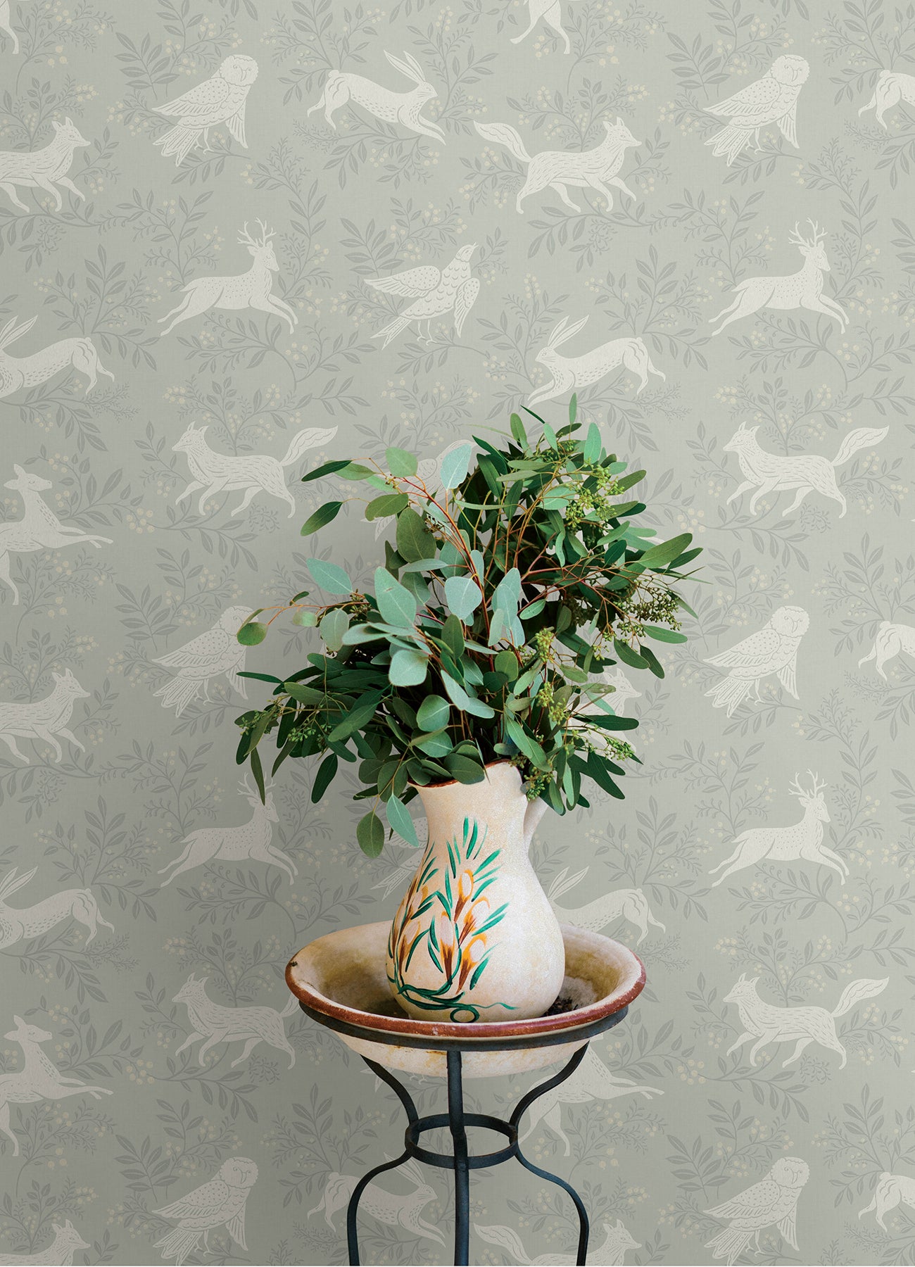 Cream ceramic vase with teal and orange motif, greens on a metal stand, set against pale gray wallpaper with white birds.