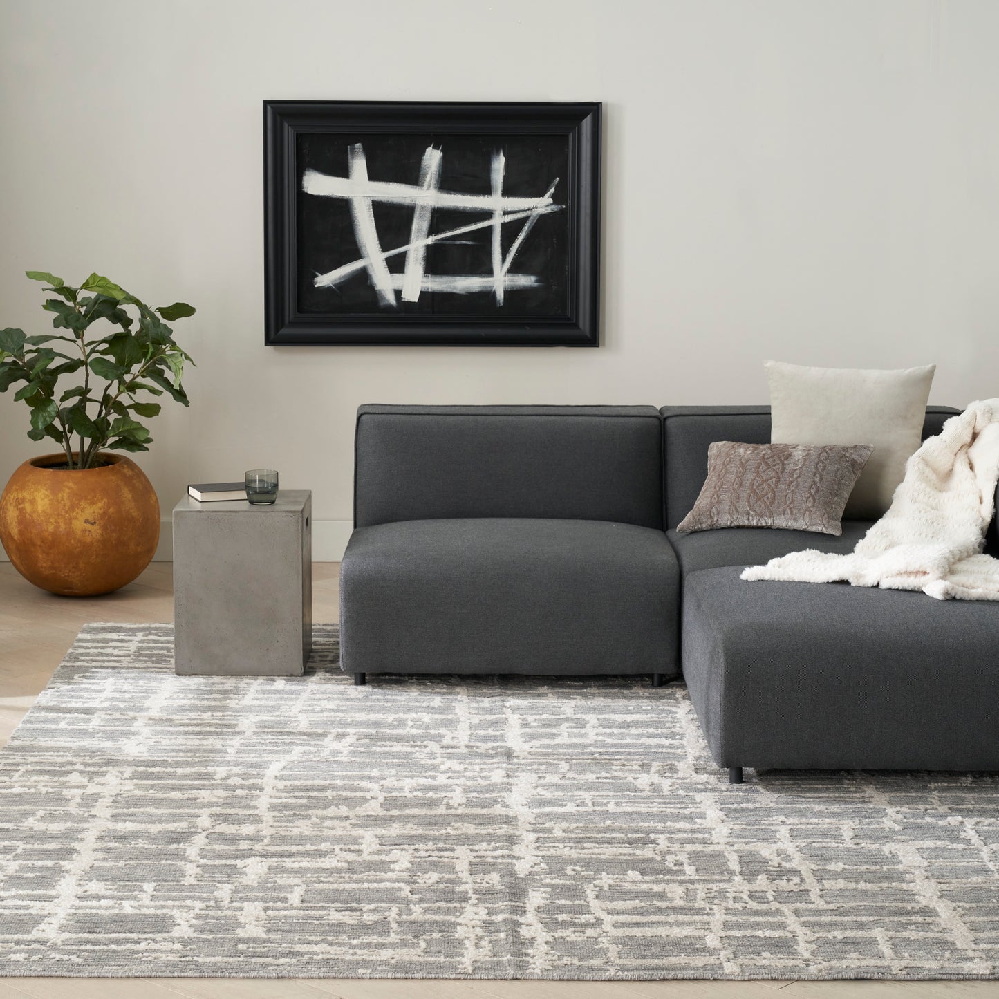 Dark gray fabric sectional with chaise, beige pillows and throw, concrete side table, geometric gray rug, black abstract art.