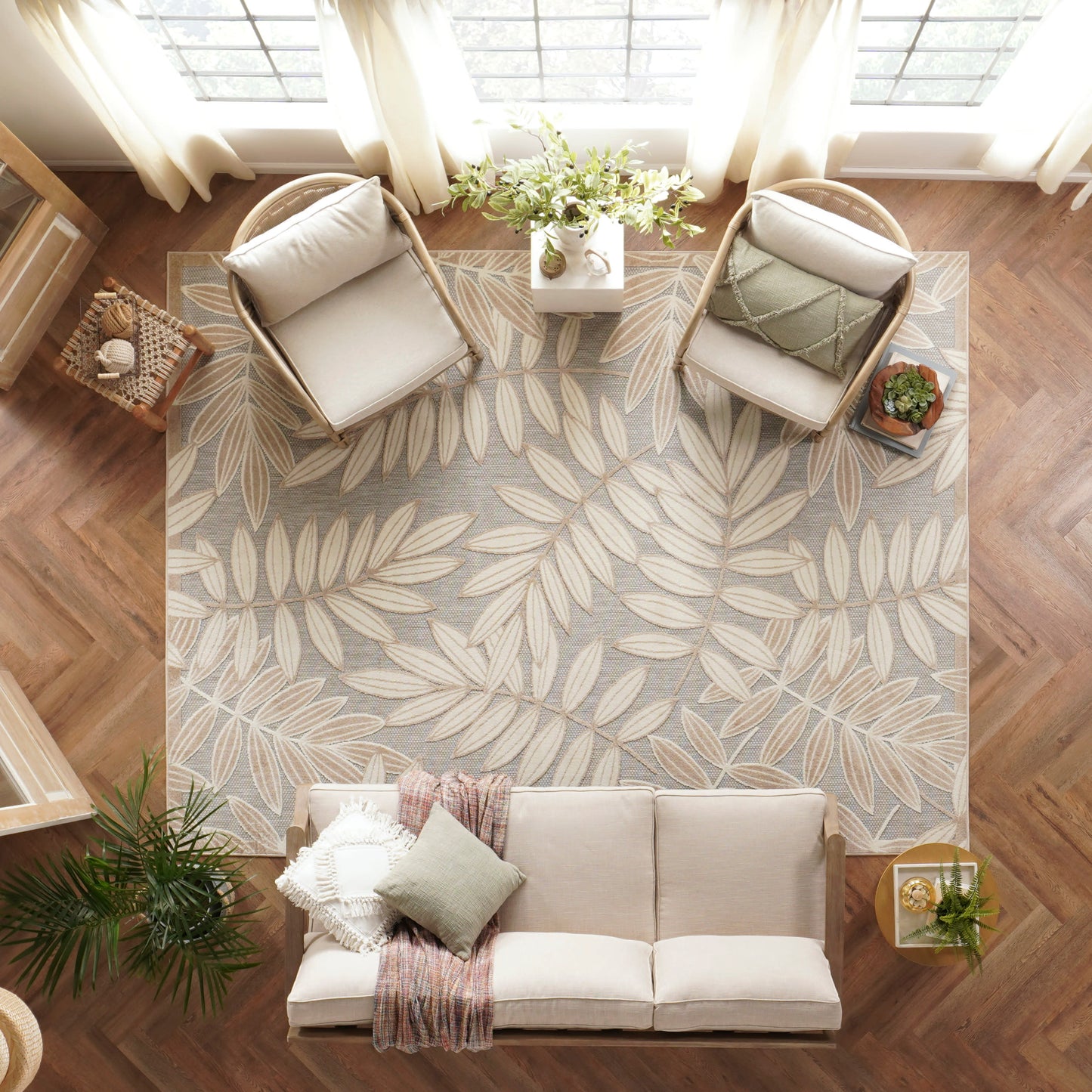 Top-down view of a cozy living room: beige sofa, two wicker chairs, gray leaf-pattern rug, wood floors, plants, and throws.