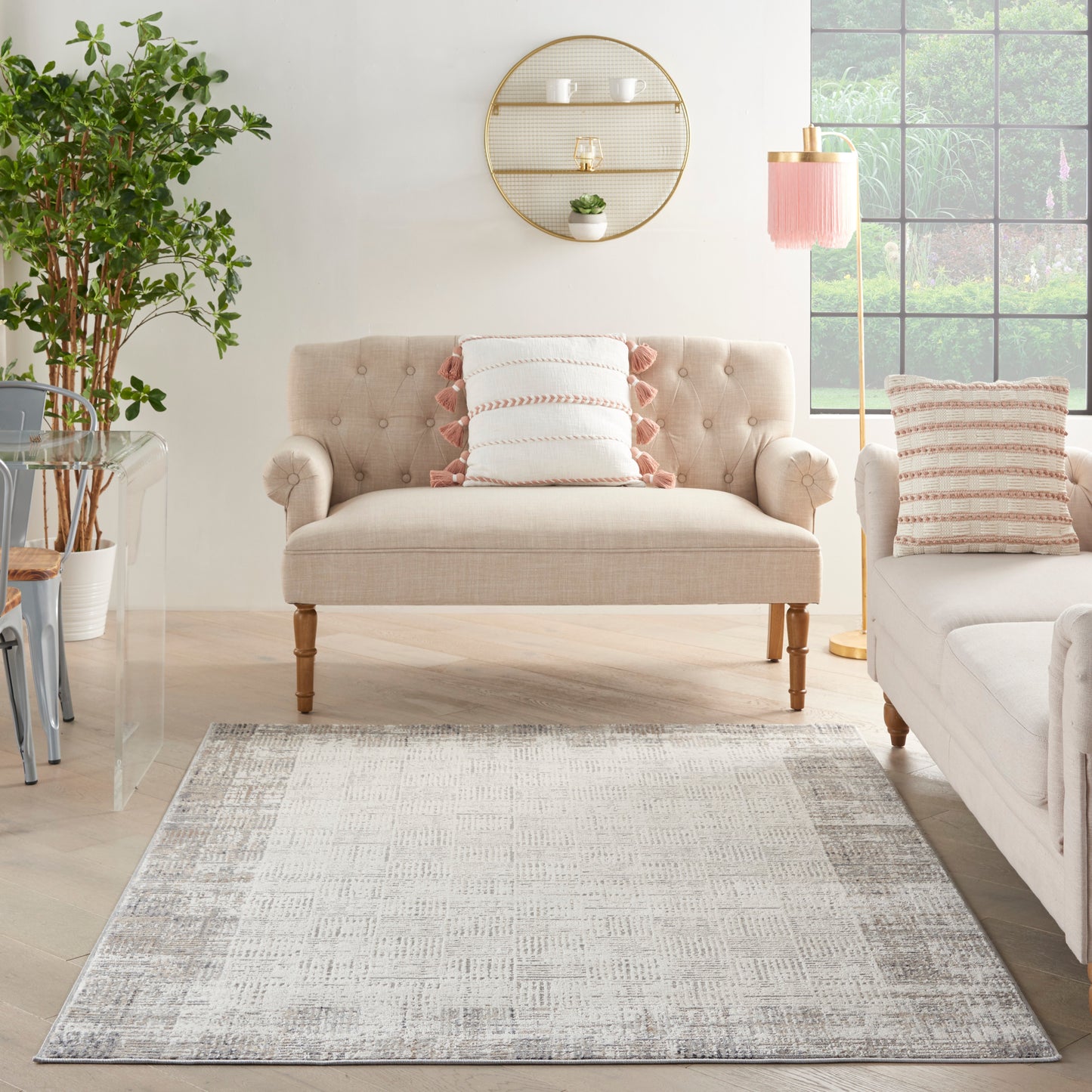 Light gray patterned rug (6' x 9') in a room with a beige tufted loveseat on wooden legs, pink fringe pillows, a shelf, pink floor lamp, and greenery.