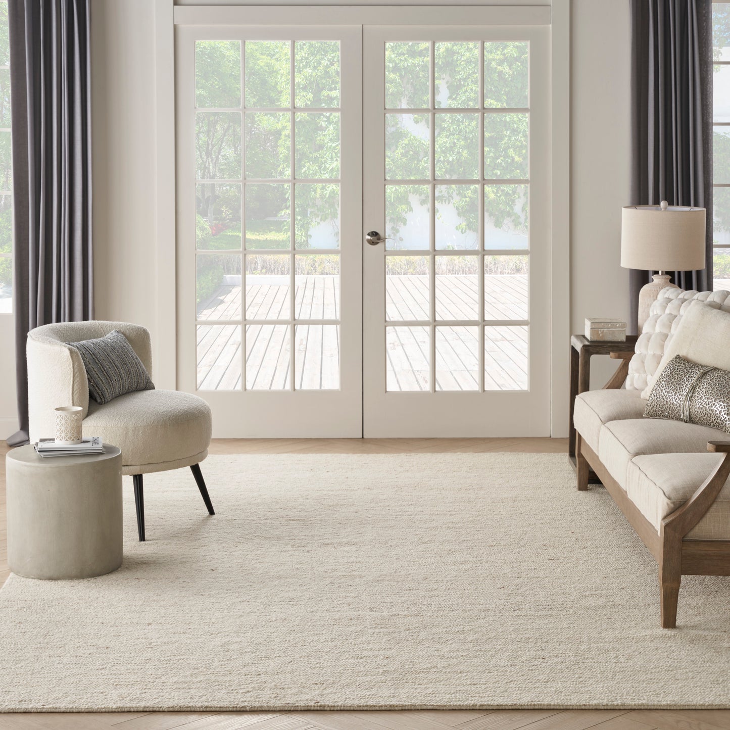 Neutral living room with beige rug, cream curved chair, beige tufted sofa, round concrete side table, wood accents & white French doors.