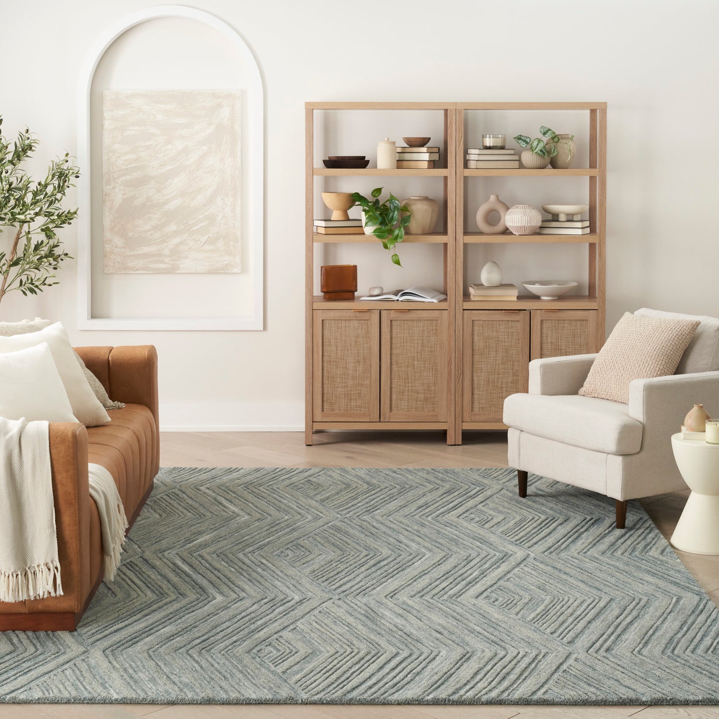 Bright living room with light wood display cabinets, blue-gray chevron rug, tan leather sofa, beige armchair, arch wall art.