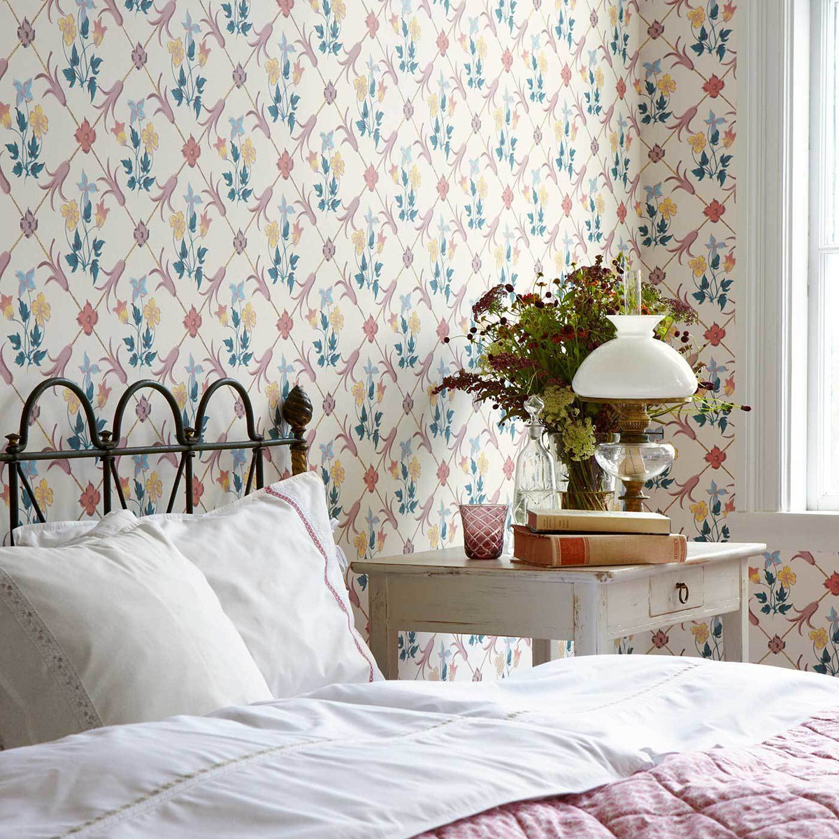 Bedroom with pastel floral wallpaper, white bedding, iron bed frame, distressed nightstand, vintage lamp, vase of flowers.