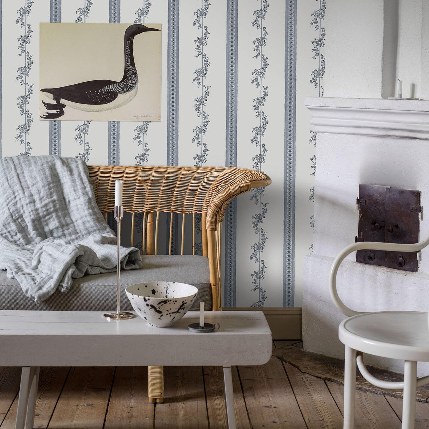 Cozy living room: curved rattan chair, gray sofa, white coffee table, blue striped wallpaper, duck art, candle and bowl.