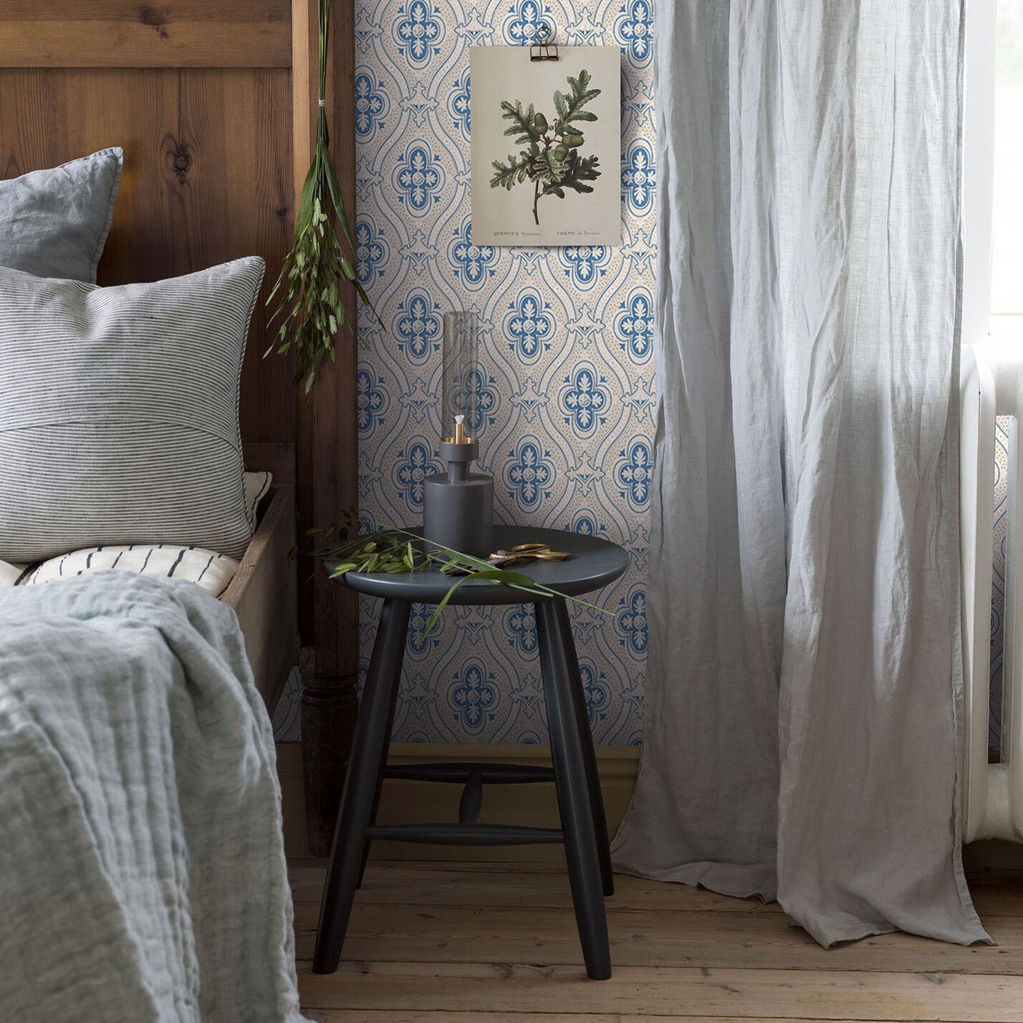 Cozy bedroom nook with wooden headboard, grey bedding, round black tripod table, plant and candle, blue wallpaper.