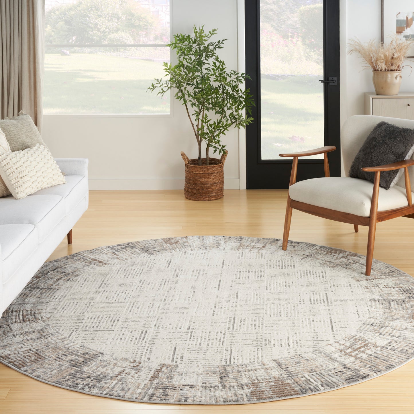 Round gray-beige patterned area rug in a bright living room with a mid-century wooden armchair, white sofa, and potted plant.