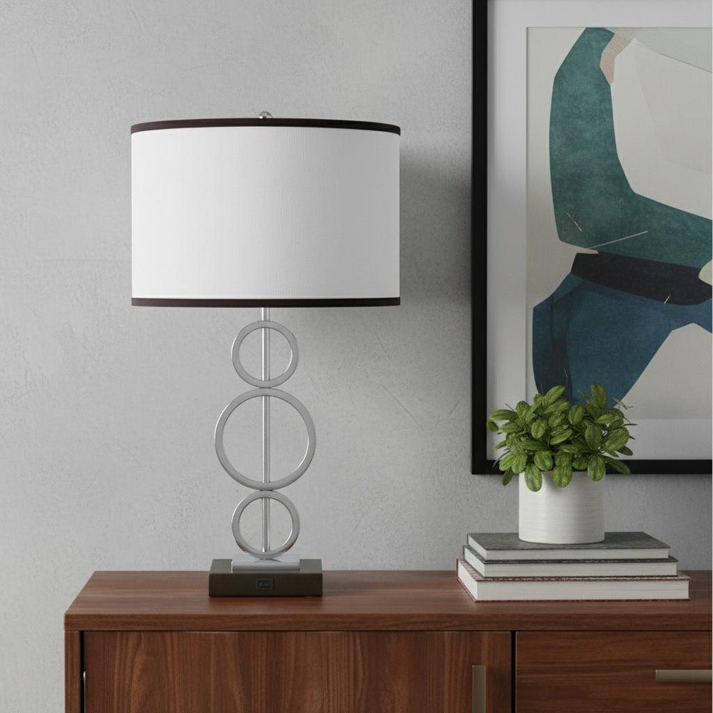 Bedside geo table lamp with white drum shade and stacked circular metal base on a bedside table beside a plant and books.