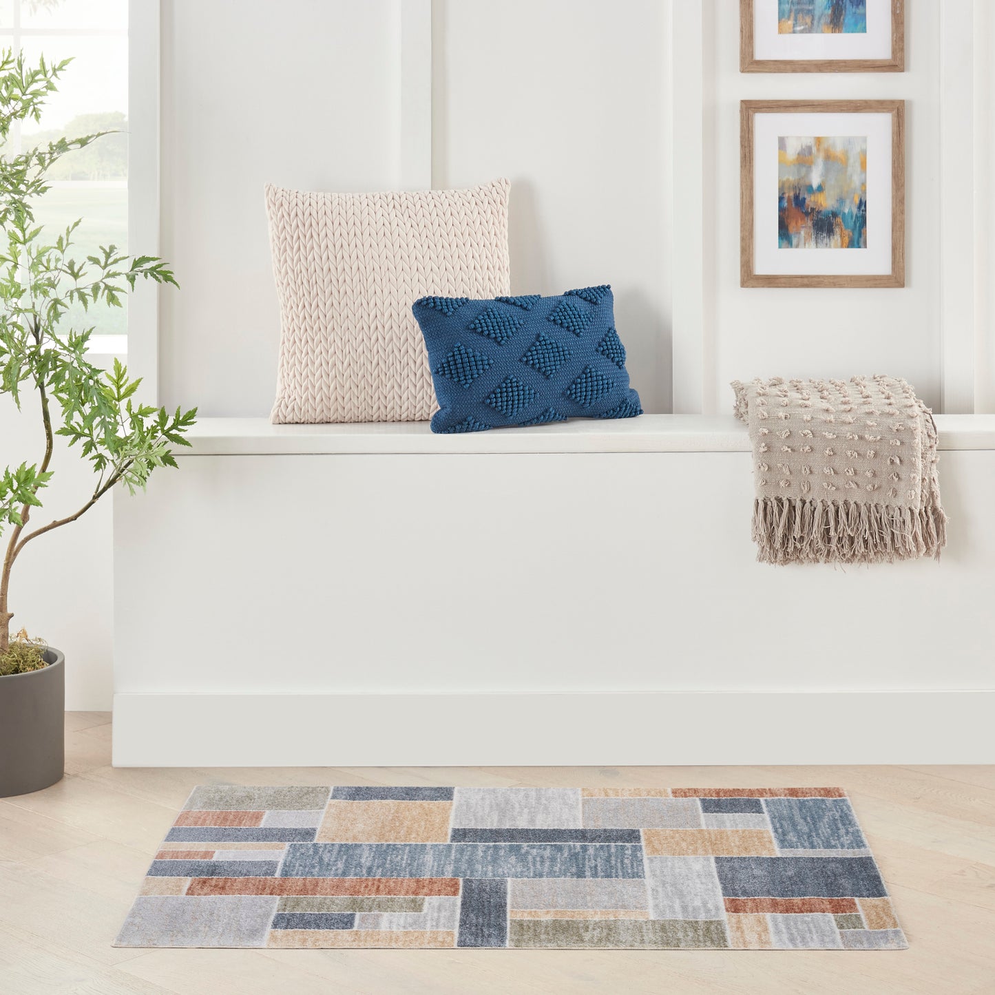 White built-in bench with beige knit pillow, blue cushion, beige fringe throw; abstract wall art, potted plant, patchwork rug.