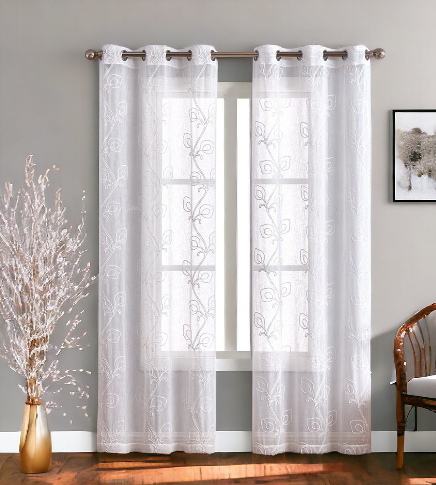 White sheer lace curtains with vine floral pattern on a metal rod with grommets, over a window in a cozy living room.