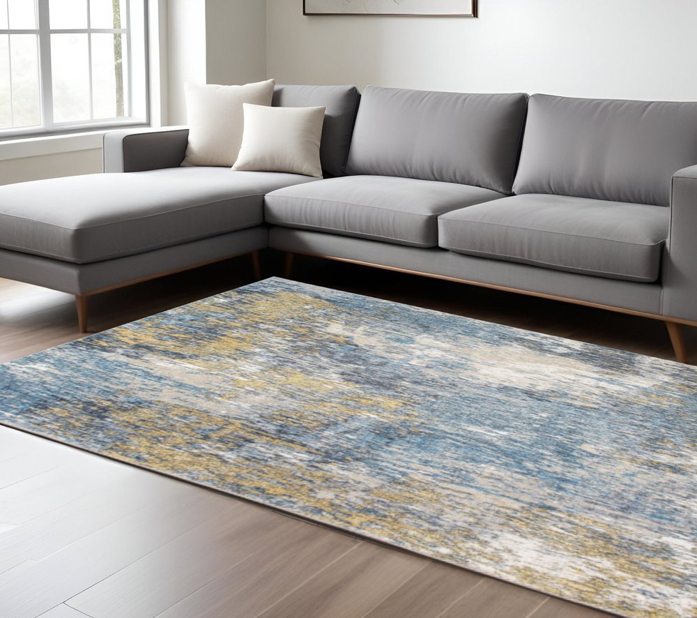 Grey L-shaped fabric sofa with wooden legs and a blue and gold abstract area rug in a bright living room.