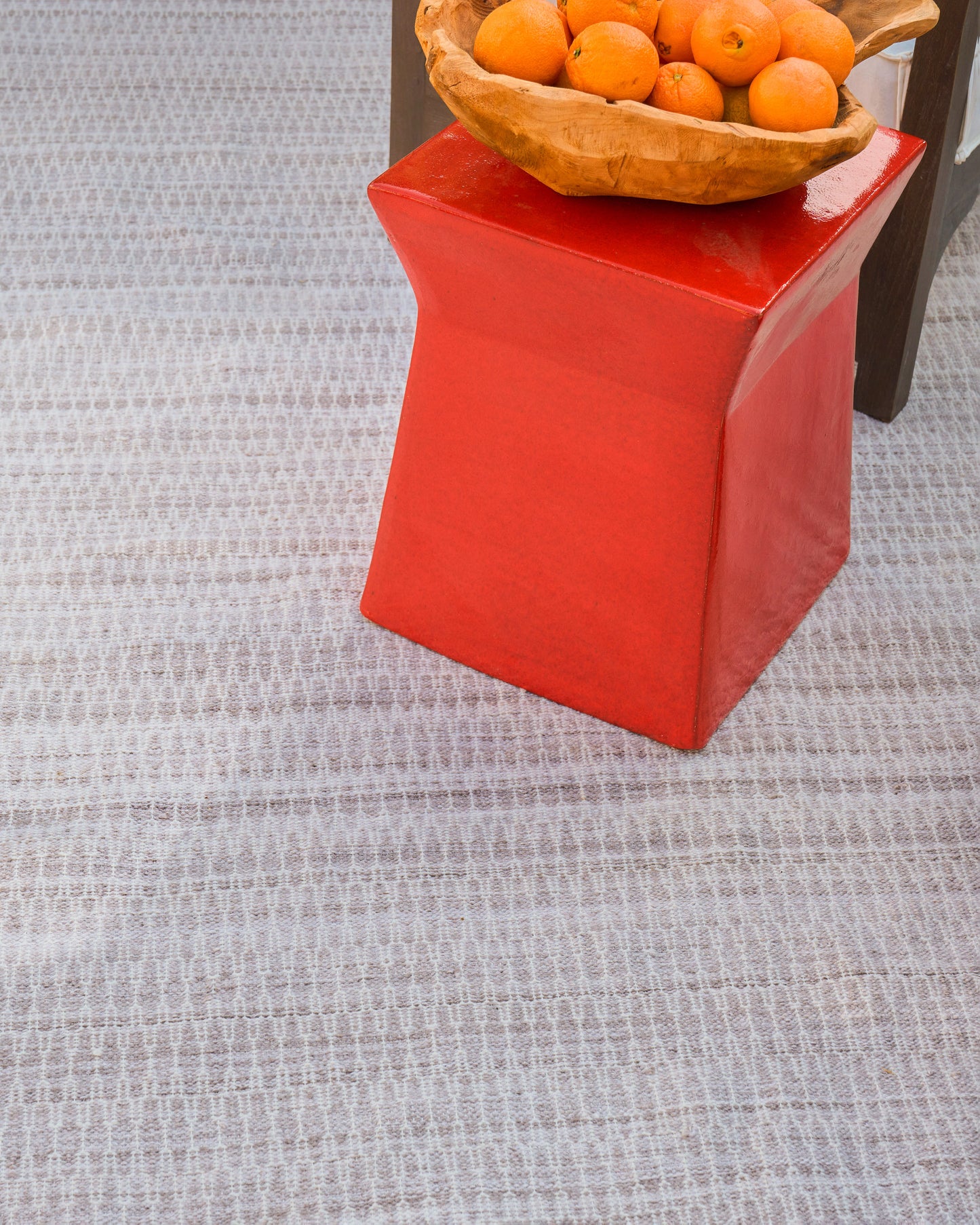 Grey flat-weave rug beneath a red curved pedestal table topped with a wooden basket of oranges.
