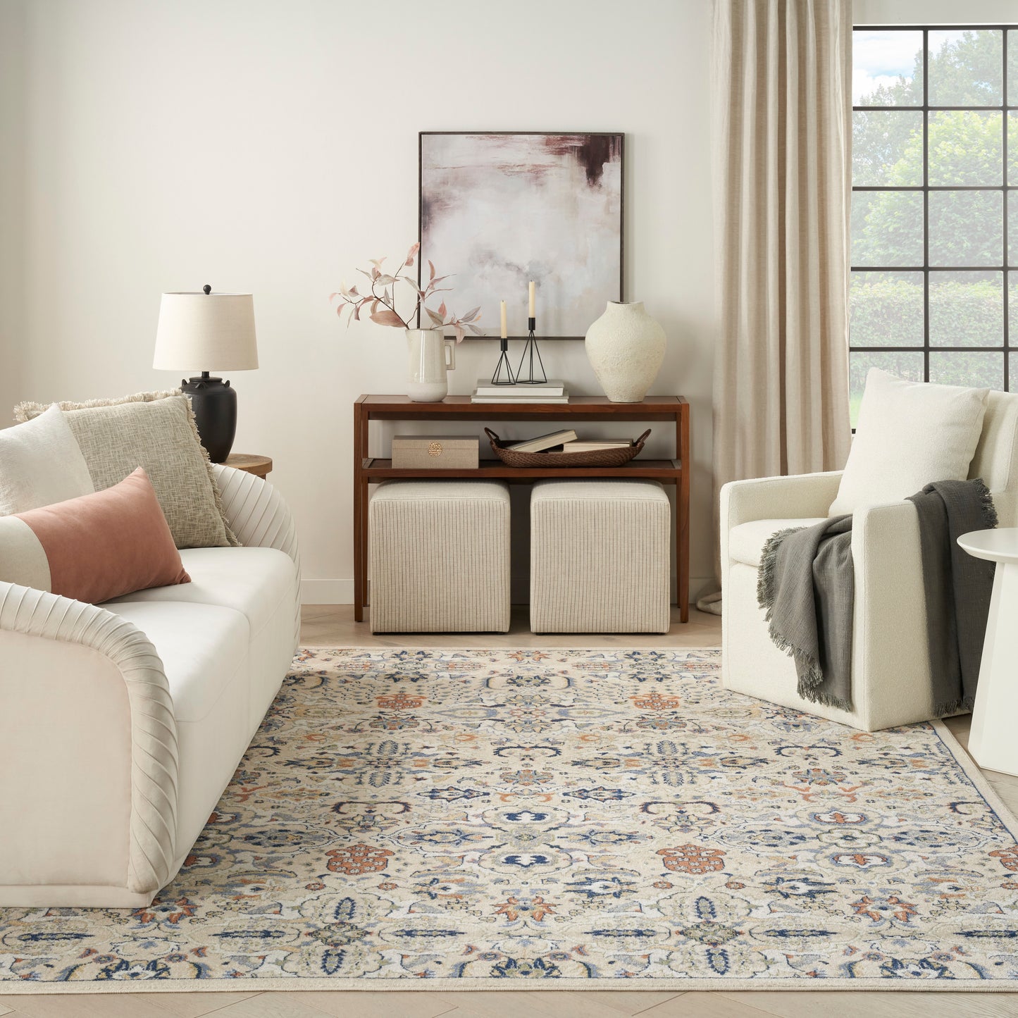 Cream upholstered sofa and armchair, beige pillows, wooden console, two beige ottomans, blue-beige patterned rug.