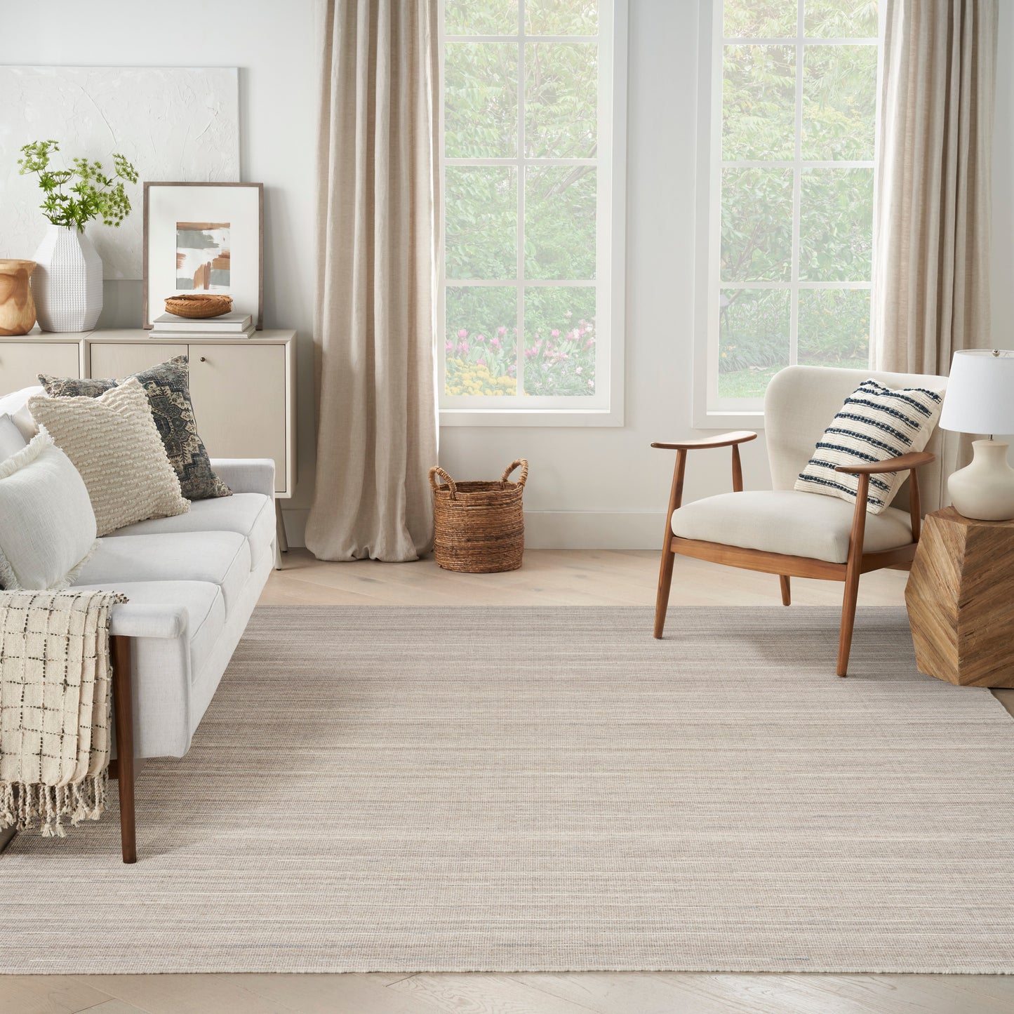 Neutral living room with light gray sofa, wooden chair, textured rug, woven basket, framed art, plant, and beige curtains.
