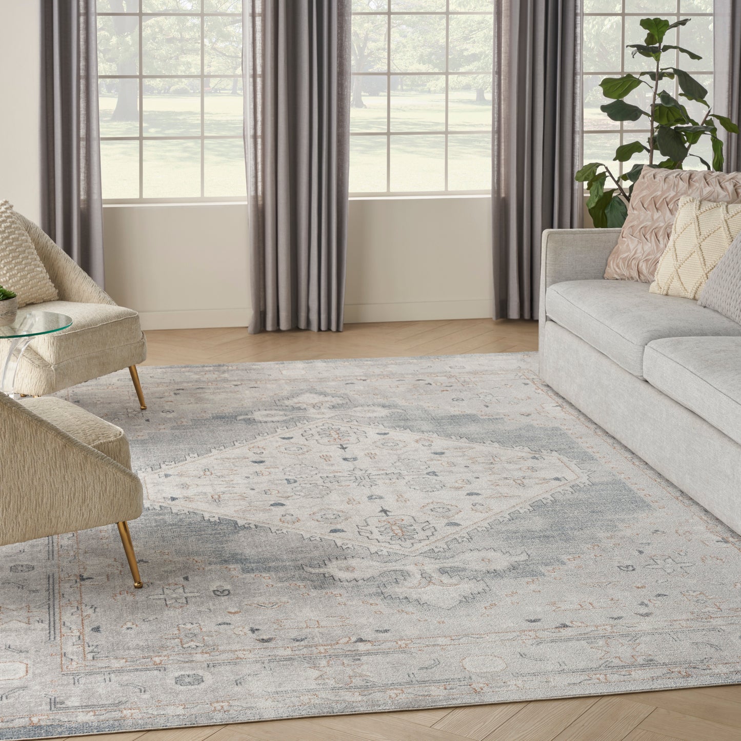 Cozy living room with light gray sofa, beige accent chairs, glass coffee table, blue-gray rug, gray curtains, and a plant.
