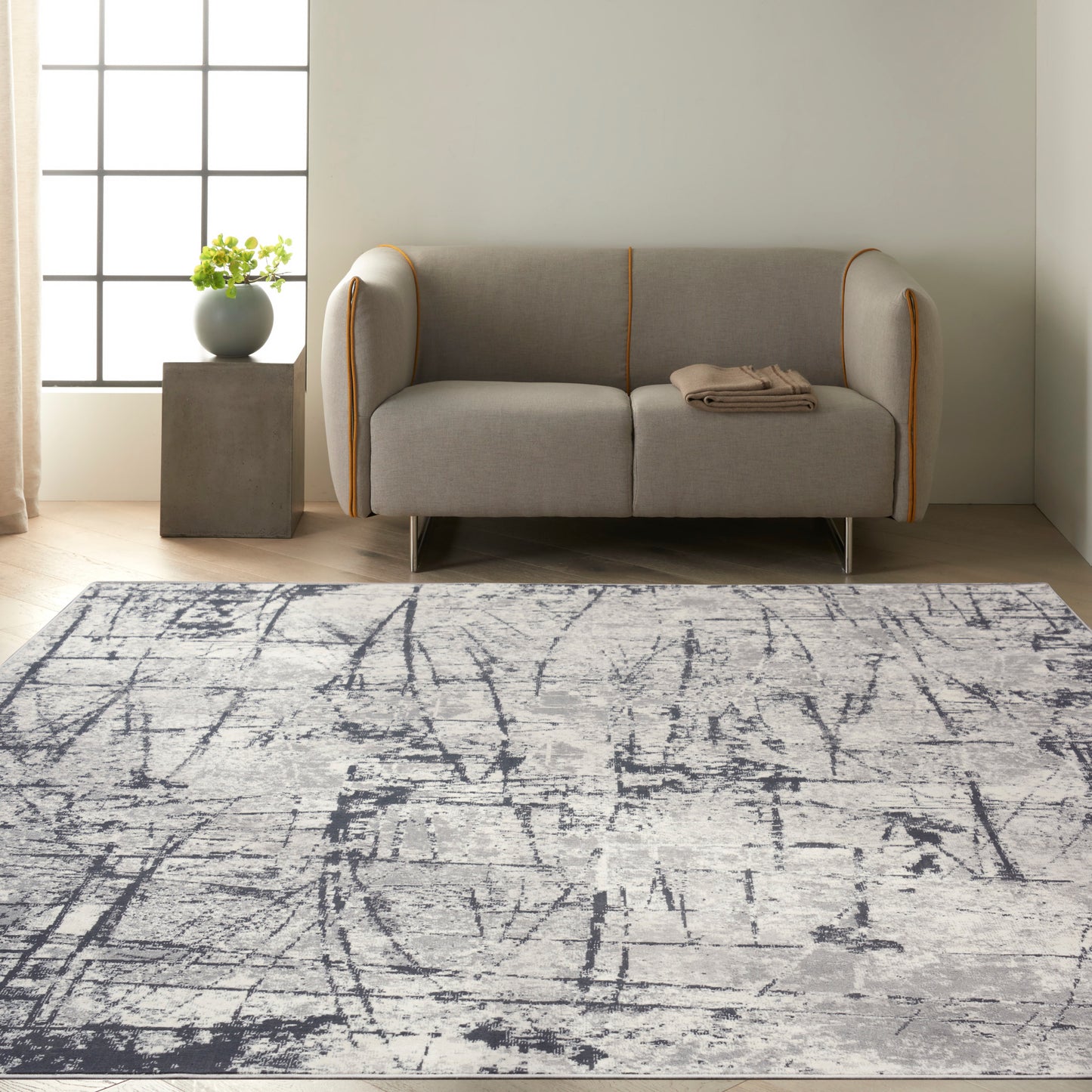 Gray two-seat sofa with orange piping and metal legs, folded beige throw; plant on pedestal by window and abstract gray rug.