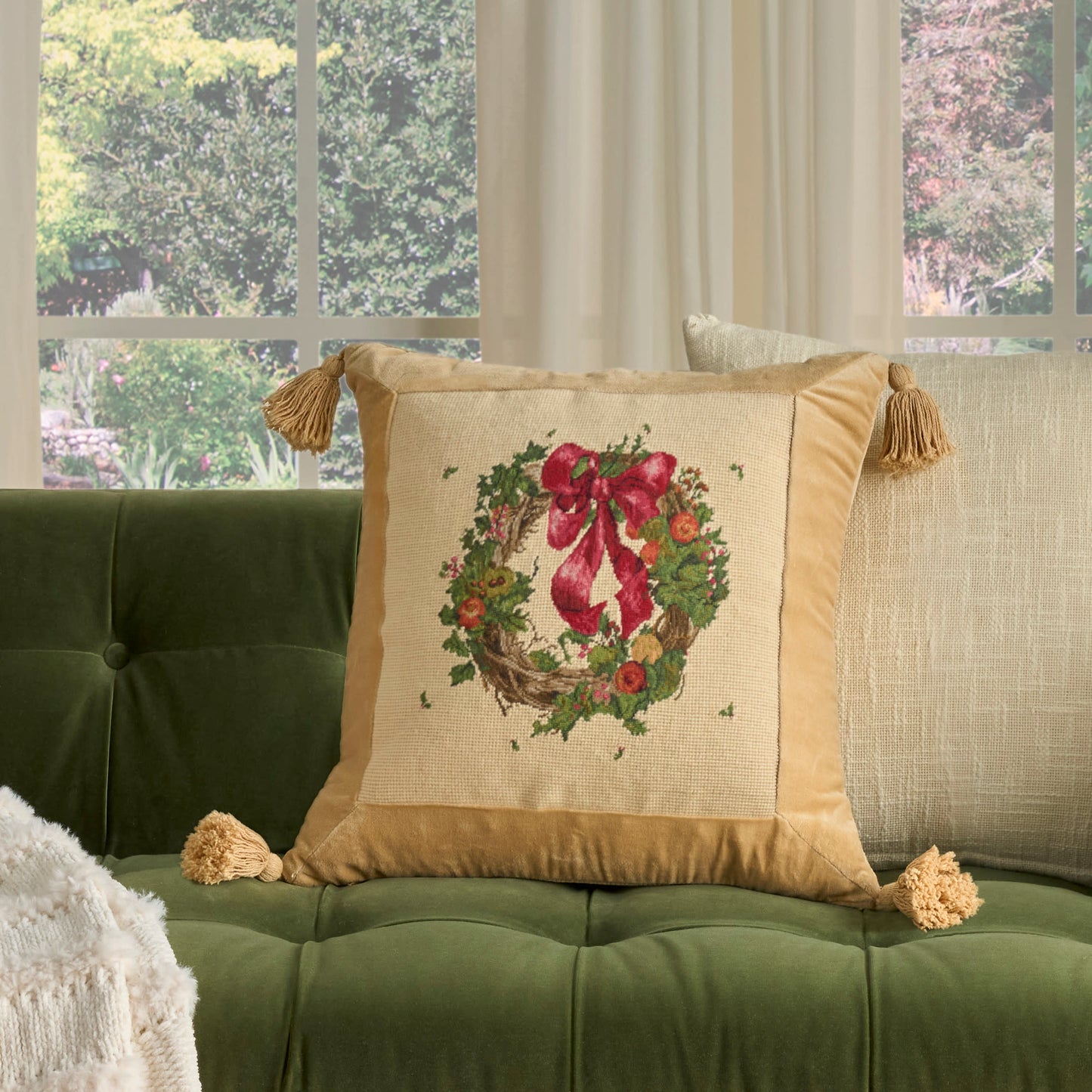 Golden-tan decorative pillow with Christmas wreath embroidery and a red bow, tassel fringes on a green tufted sofa.