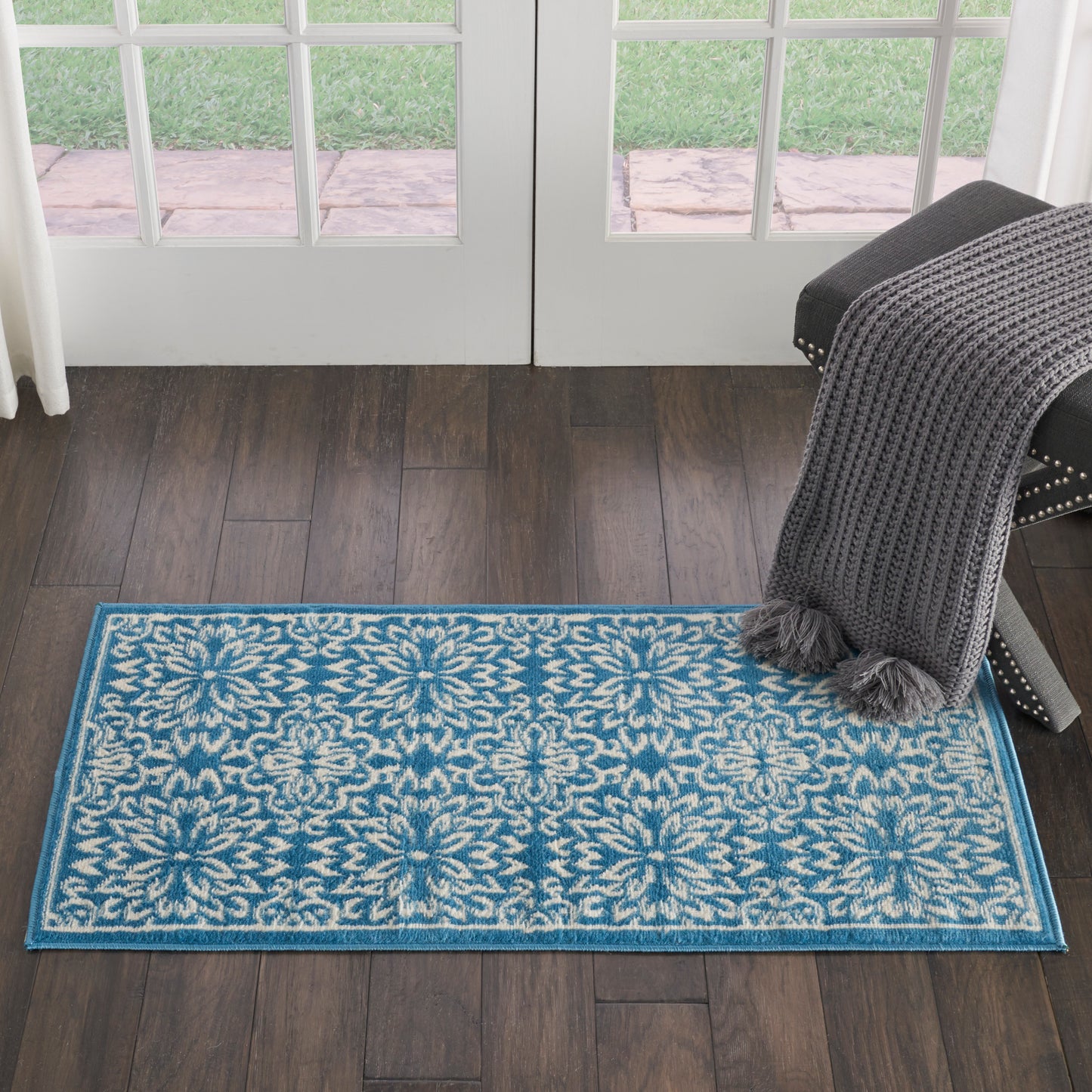 Rectangular teal-blue area rug with white damask floral pattern and blue border; draped dark gray knit throw on chair nearby.