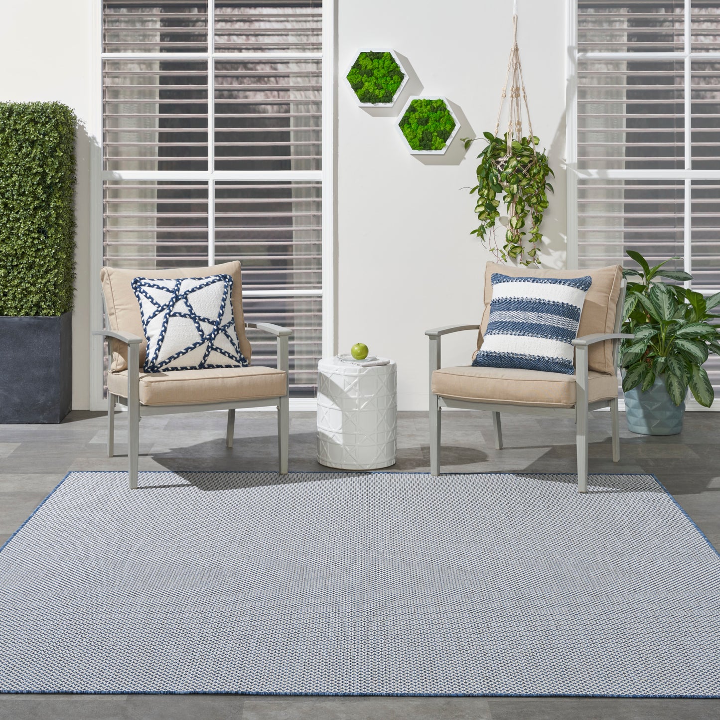 Modern outdoor seating with beige cushioned metal chairs, blue-white pillows, white side table, gray rug with blue border.