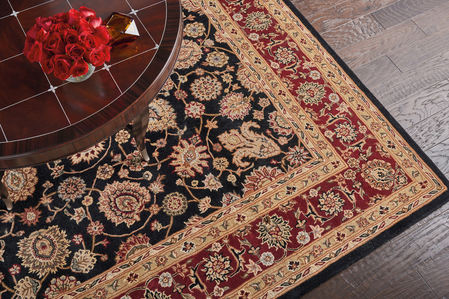 Persian-style oriental rug with black field, gold floral motifs and red border; round wooden table with red roses.