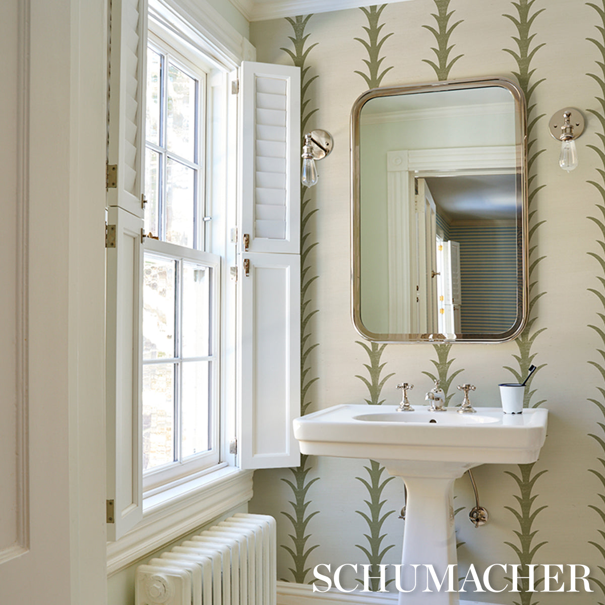 Bathroom scene with olive acanthus stripe wallpaper, white pedestal sink with chrome faucets, oval-framed mirror, and a window with shutters.