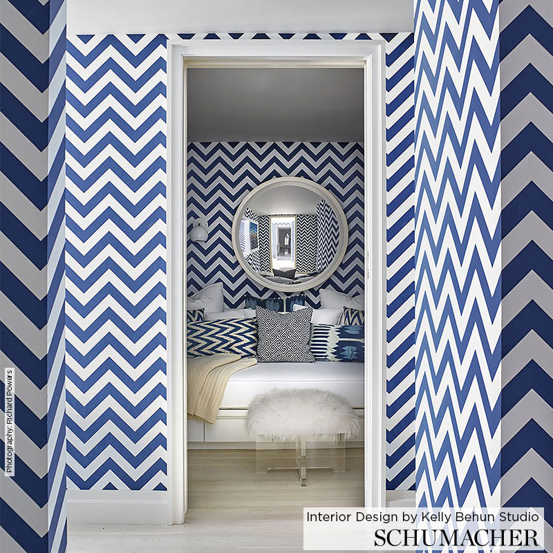 Blue and white chevron wallpaper across walls; round wall mirror, white bed with blue pillows, fluffy white ottoman.