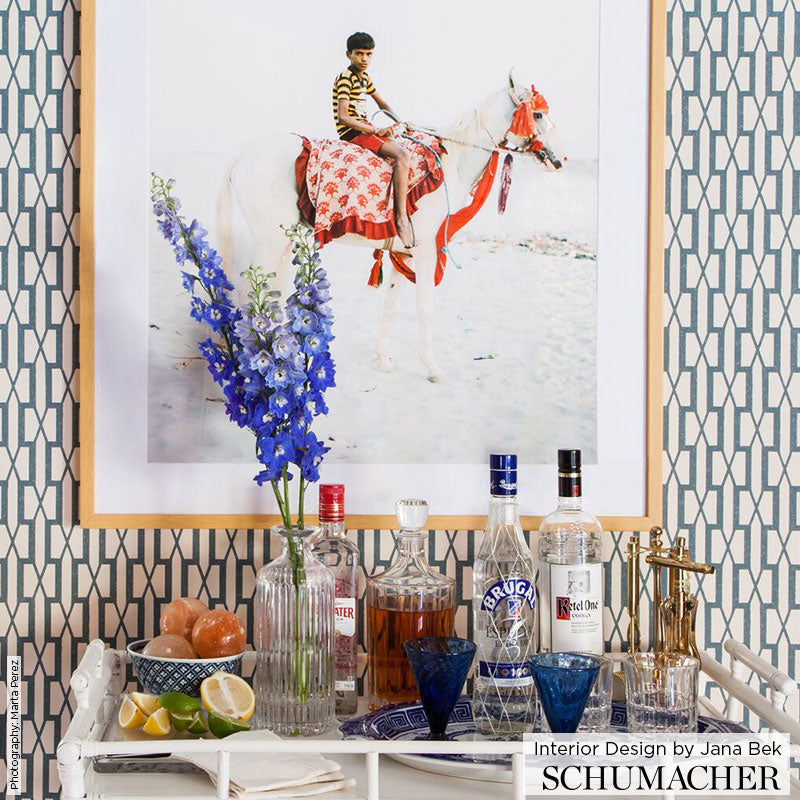 White bar cart with glass liquor bottles, blue stemware, lemons, tall vase of blue delphiniums, framed horse artwork.