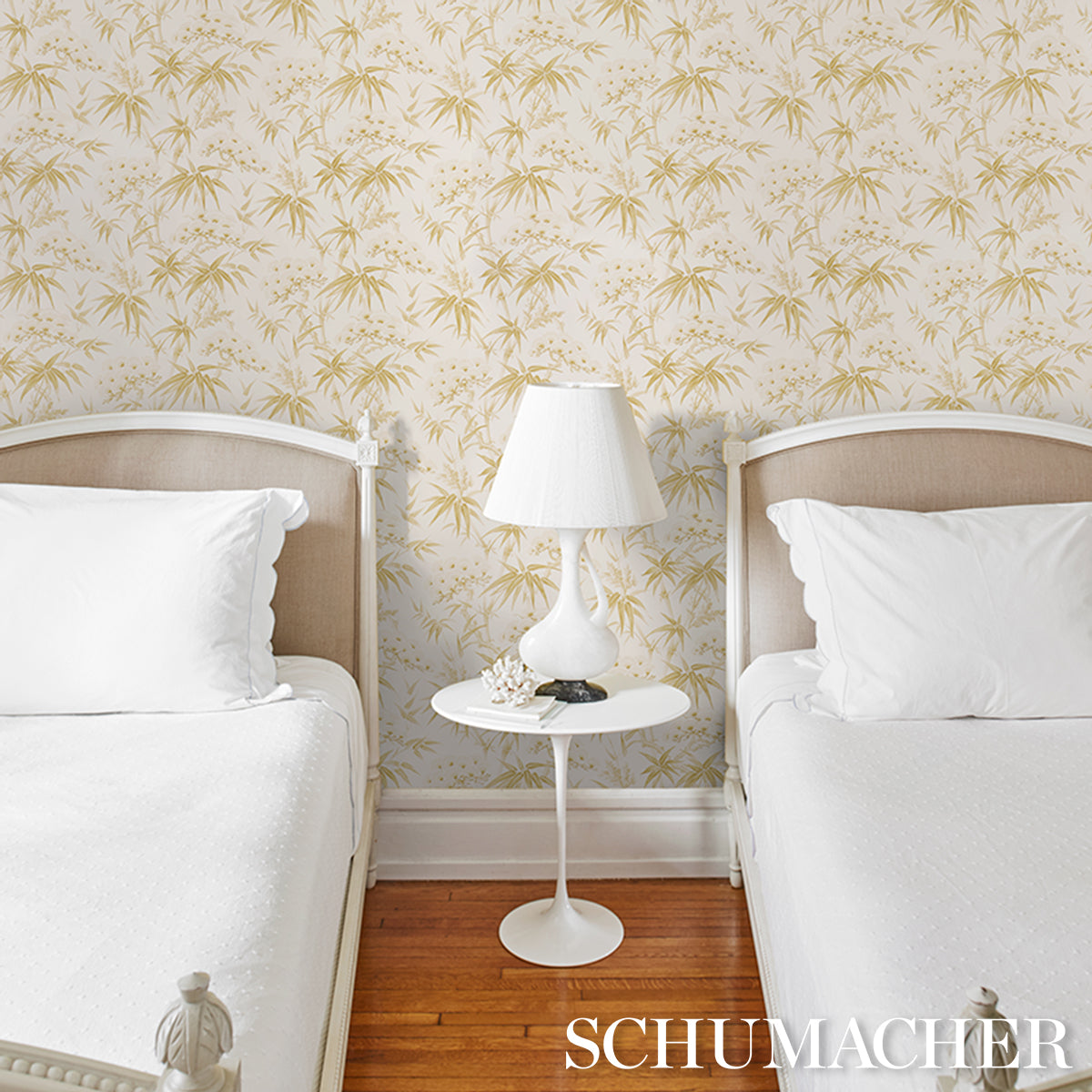 Twin beds with white linens, round white pedestal nightstand, ceramic lamp; palm-leaf wallpaper and wood floor.