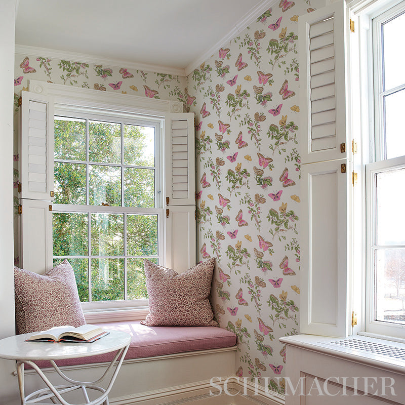 Sunlit window seat with pink cushioned bench, two pink pillows, round white table with book, butterfly-floral wallpaper.