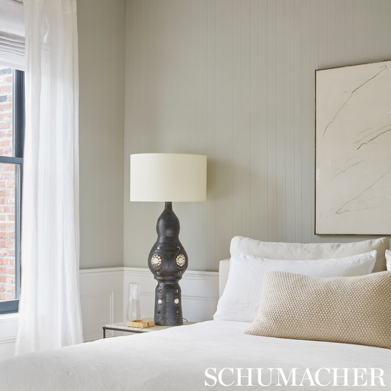 Neutral bedroom scene with white bedding, beige pillow, black ceramic lamp, glass carafe on side table, gray paneled walls.