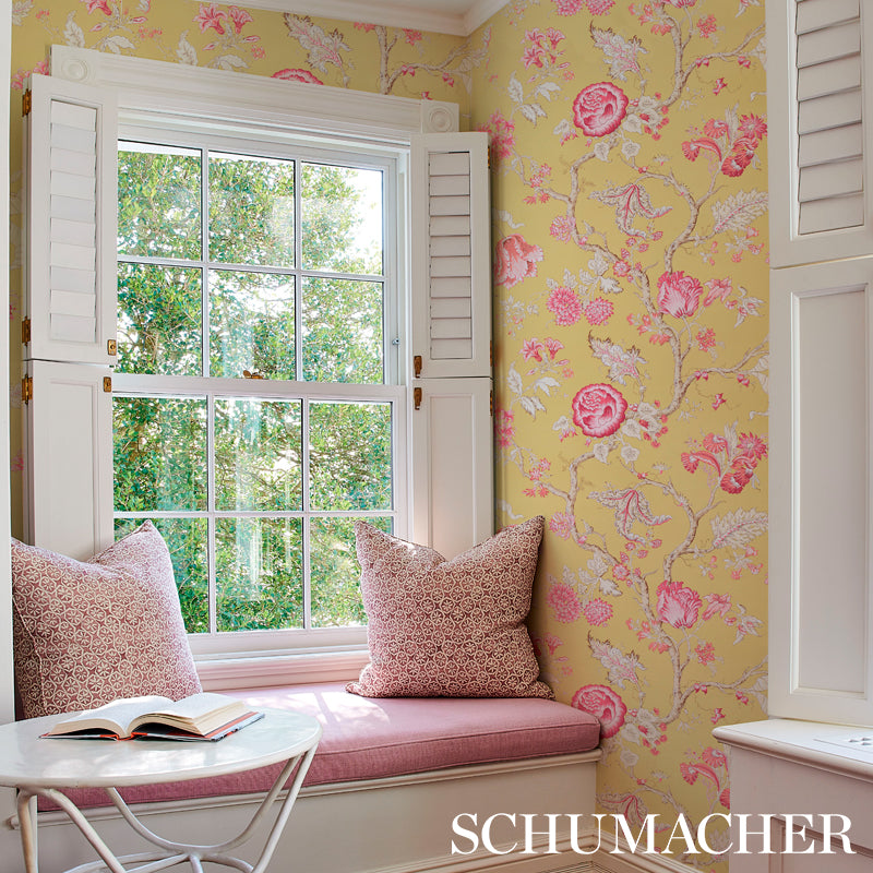 Built-in white window seat with pink cushions, two pillows, blue floral wallpaper, small white round table with open book.