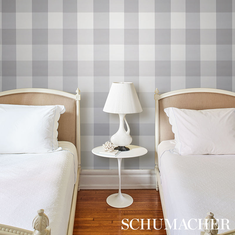 Gray plaid wallcovering with large checks visible behind a bedroom setup featuring two twin beds with beige headboards, white bedding, and a round white bedside table with a lamp between them.