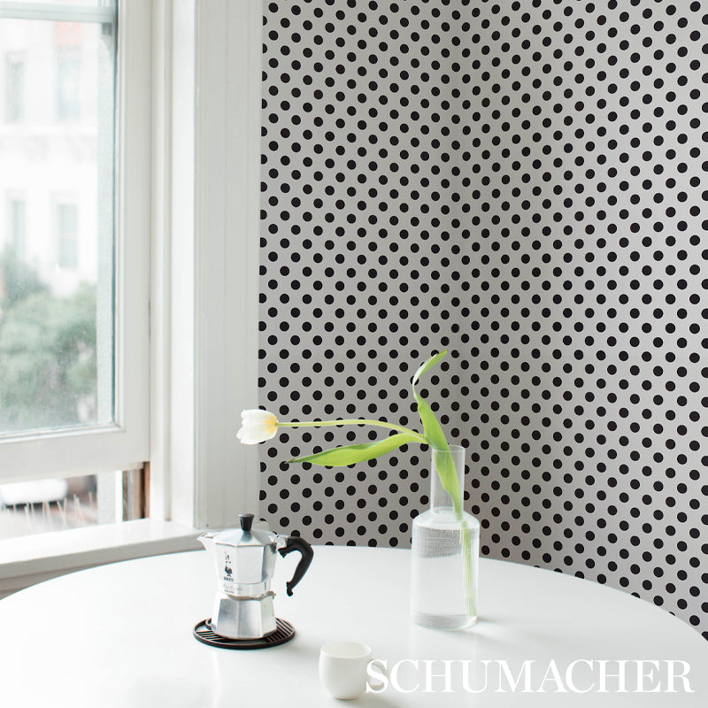 Moka pot in stainless steel on a round white table beside a glass bottle with a white tulip, against black polka-dot wallpaper.